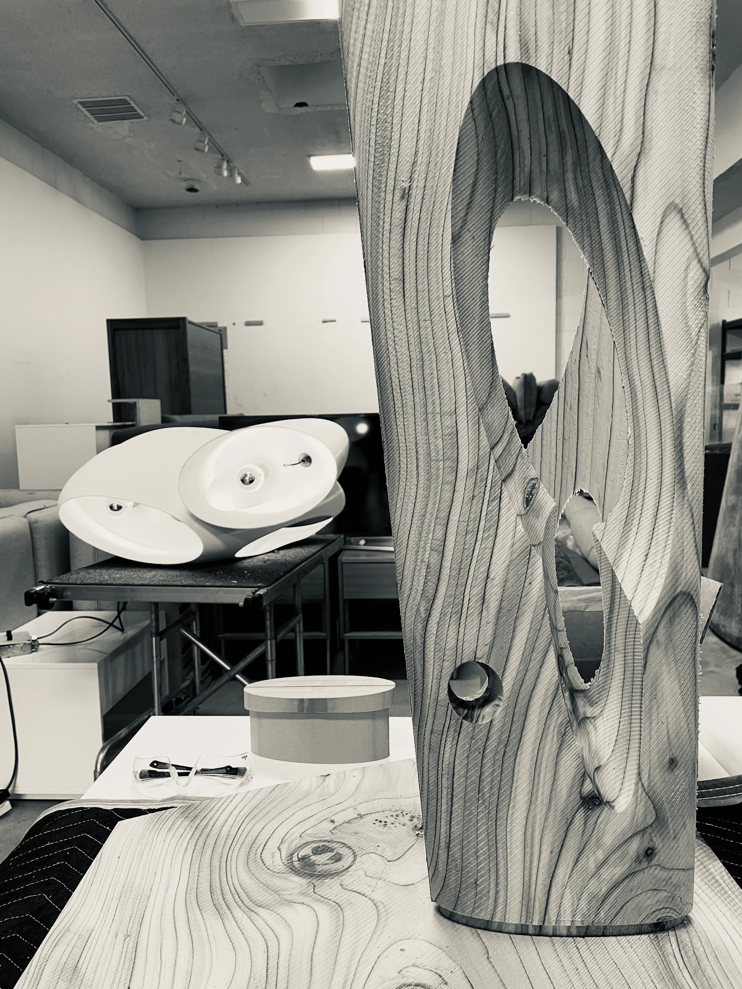 A wooden sculpture in an art studio surrounded by furniture and art supplies, with a white gallery / workshopbackground.