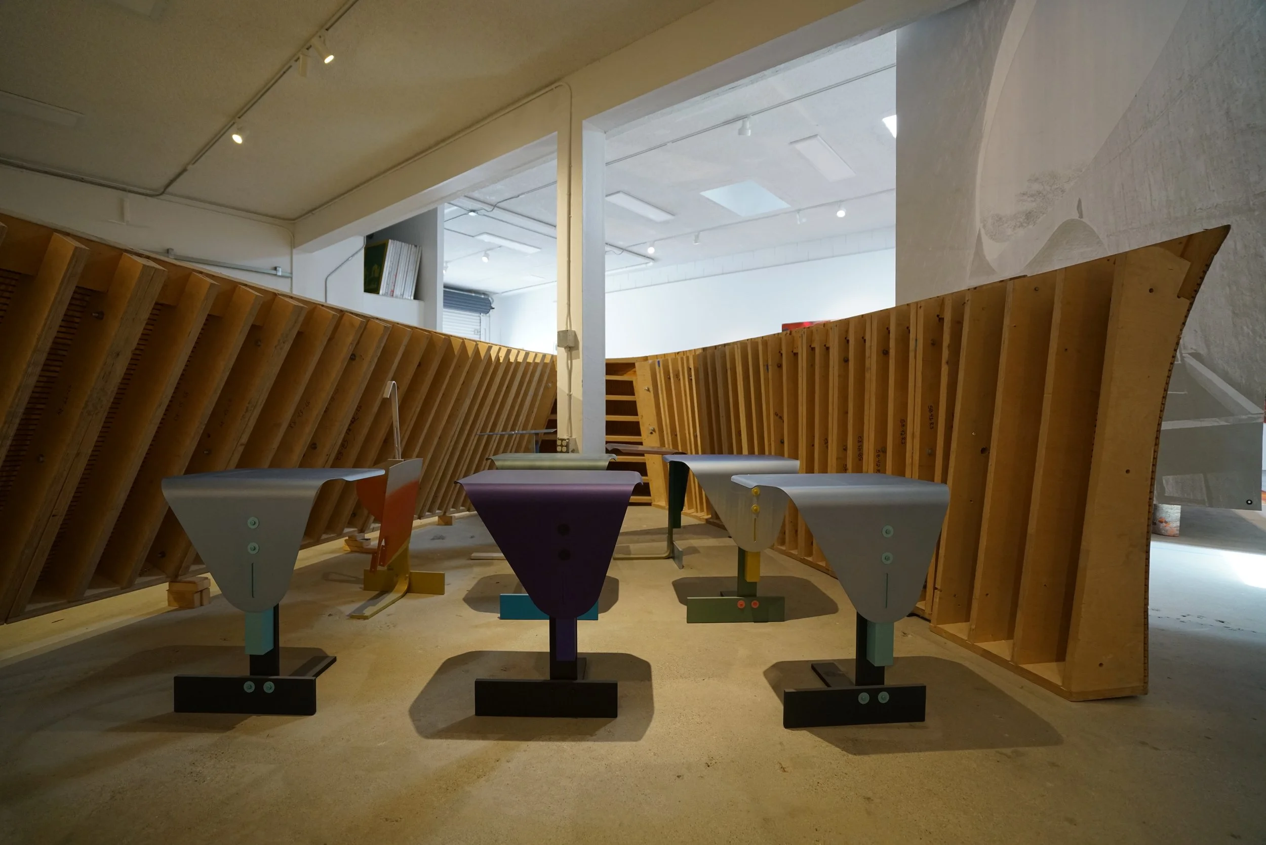 Interior view of an art installation featuring colorful geometric shapes and wooden structures with angled, slanted walls in an exhibition space.