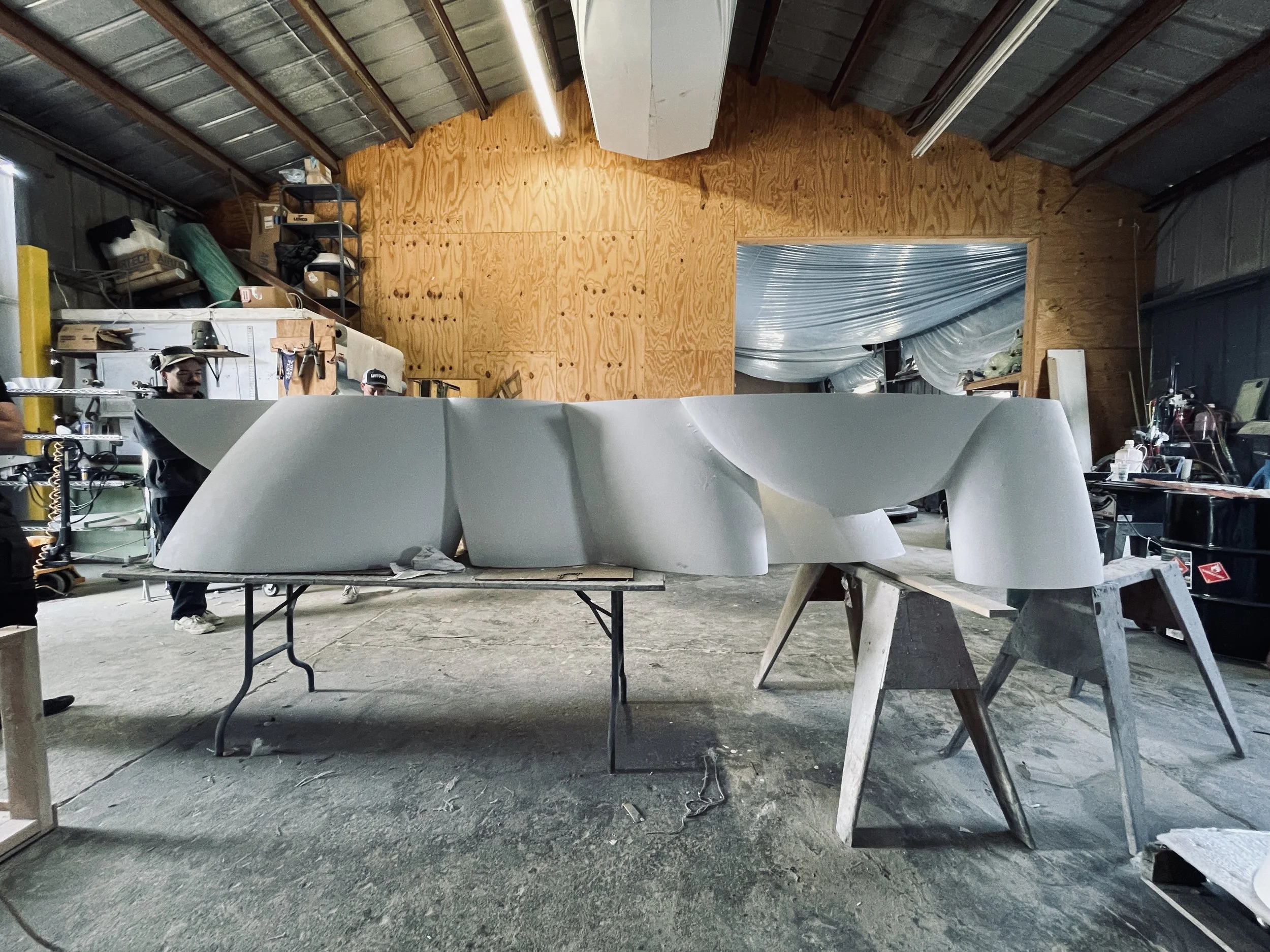 Workshop with large curved white fiberglass sculpture, in progress, surrounded by tools, workbenches, and shelves.