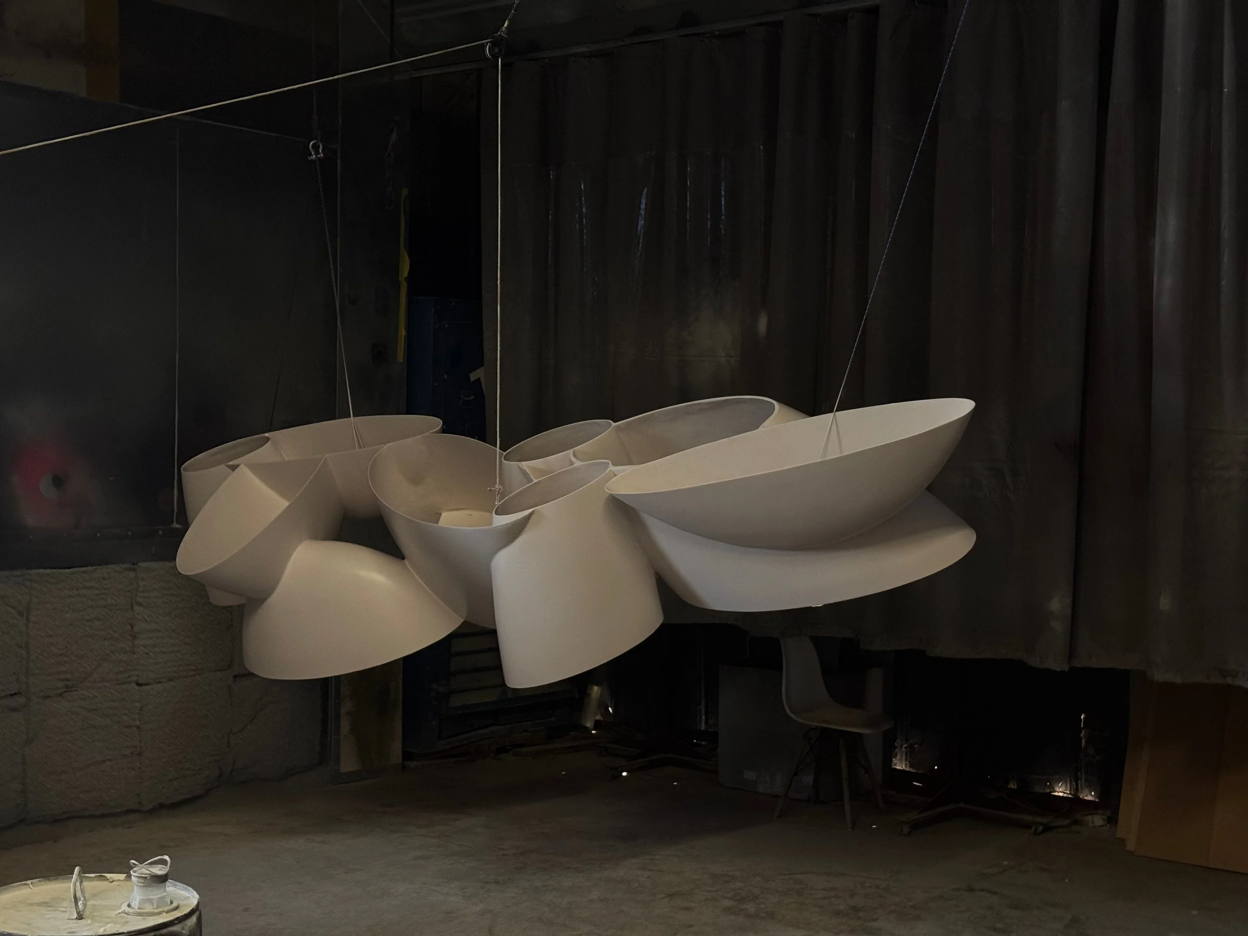 White fiberglass sculpture in construction, with abstract curved forms suspended with thin wires in a dark studio or workshop environment.