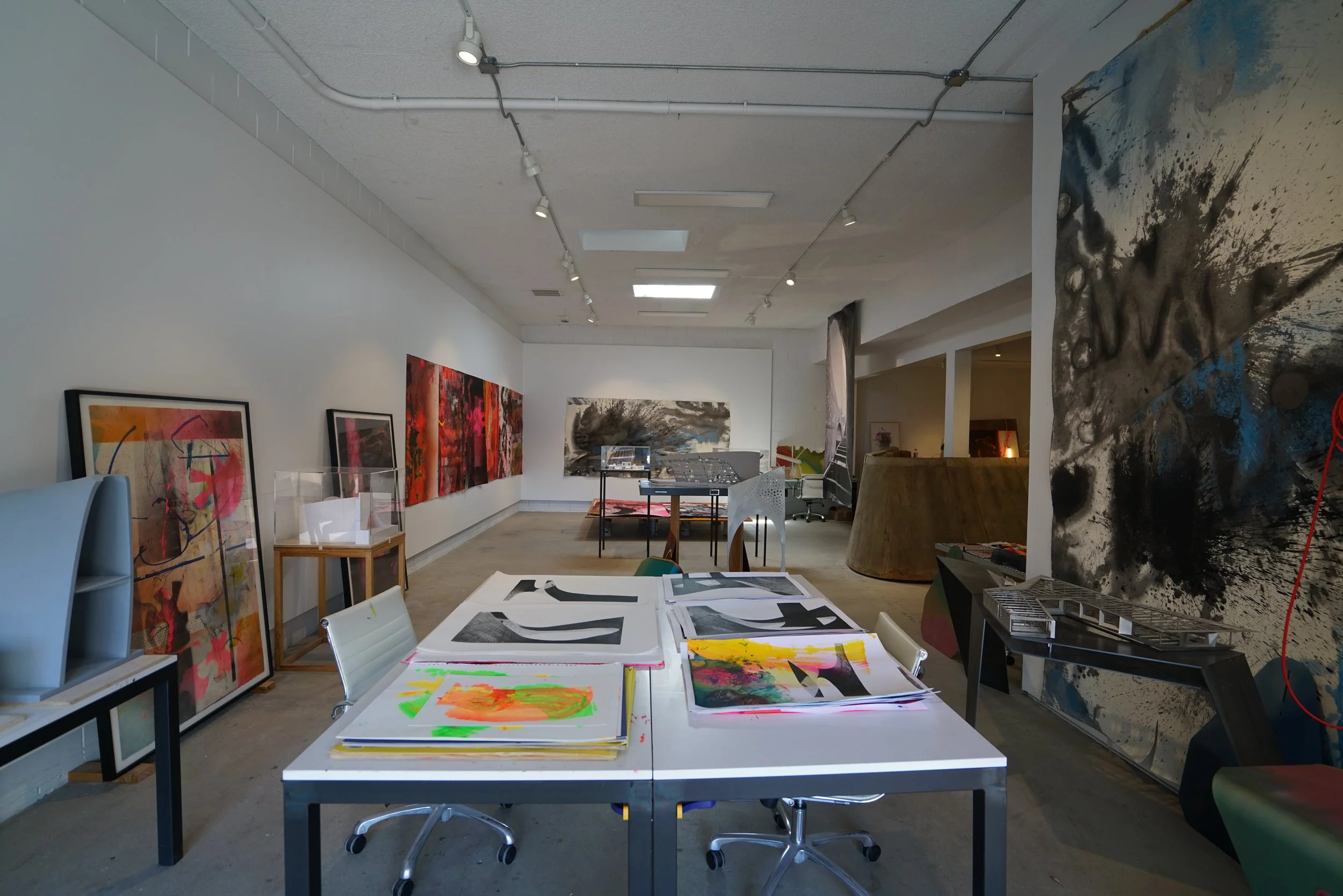 Art gallery or studio with various paintings and abstract artwork displayed on the walls and on tables. The space features white walls, track lighting on the ceiling, and a concrete floor.