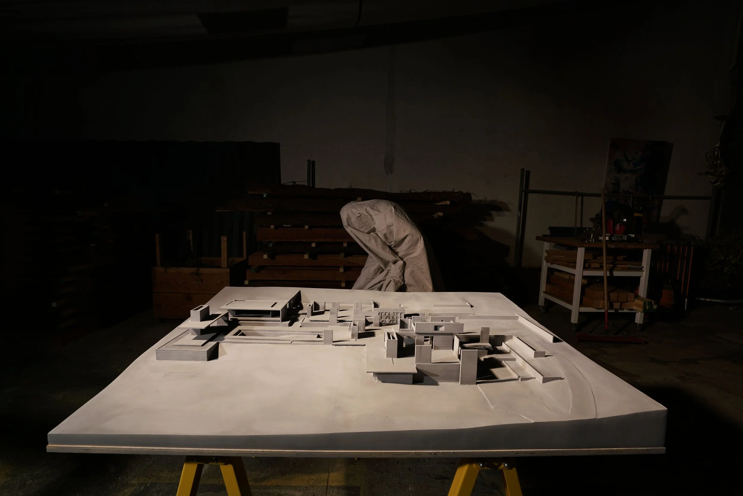 Architectural model of a building on a table in a workshop, with an abstract figure in the background.