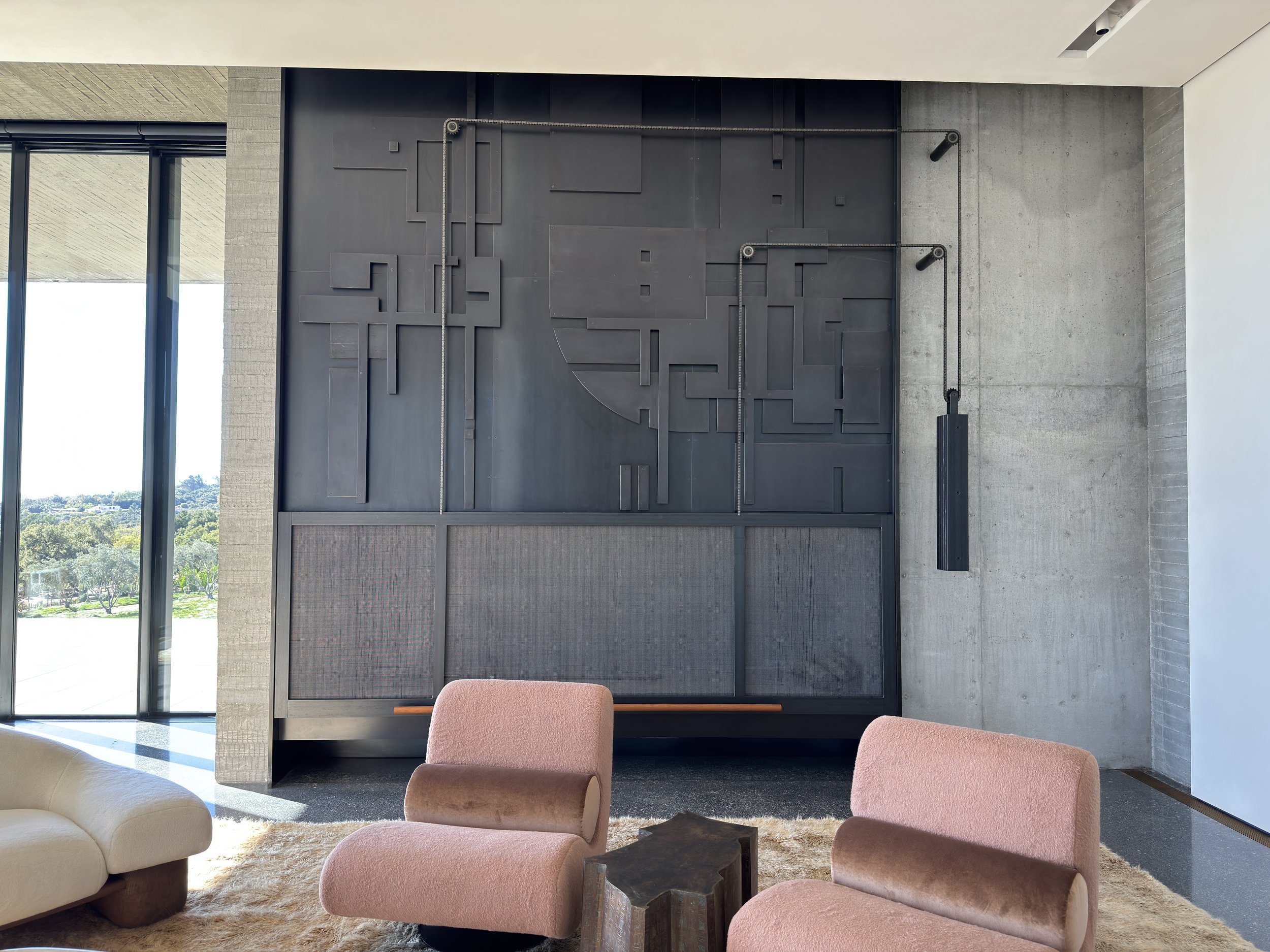 Modern interior with pink chairs, sofa, a steel geometric fireplace wall art piece on a concrete wall with large windows.