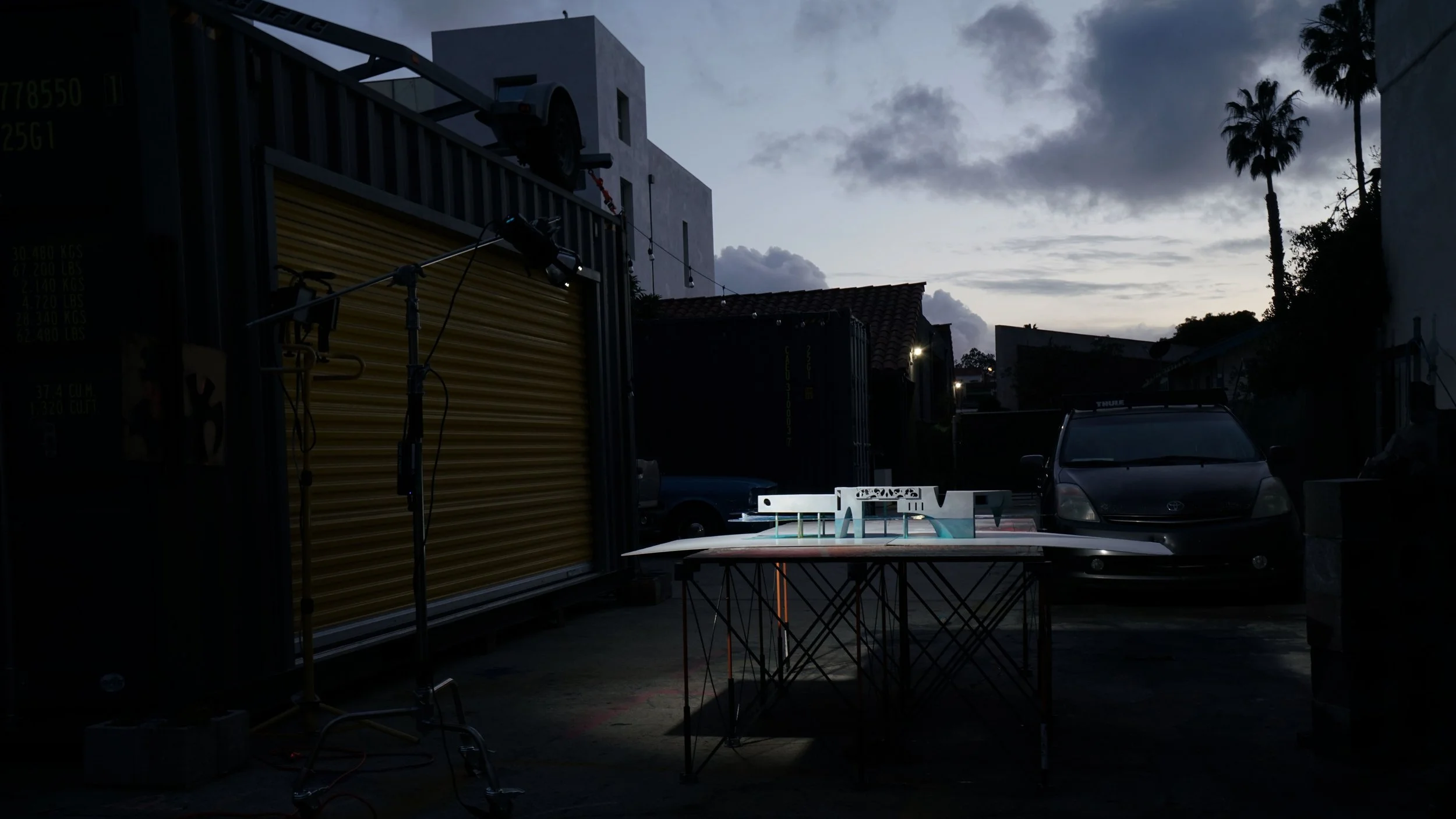 An abstract outdoor scene at dusk featuring a table with architectural models, a lighting setup with stands,  with palm trees and buildings silhouetted against a cloudy sky.