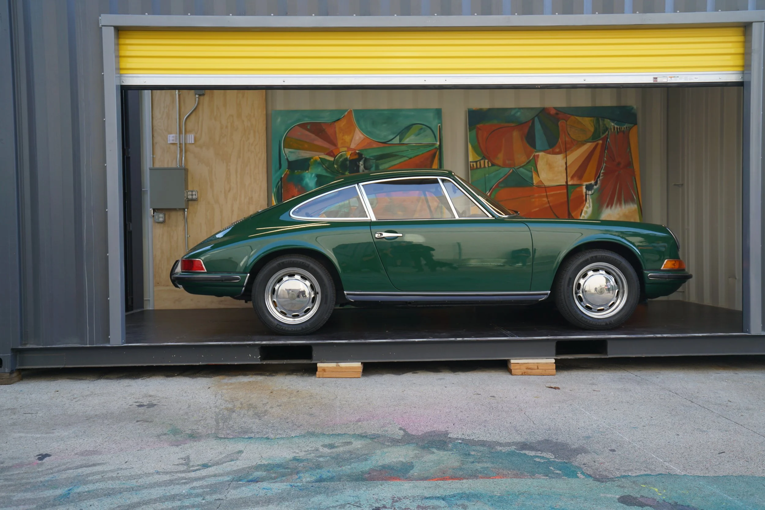 A green vintage Porsche car inside a storage container with paintings in the background.