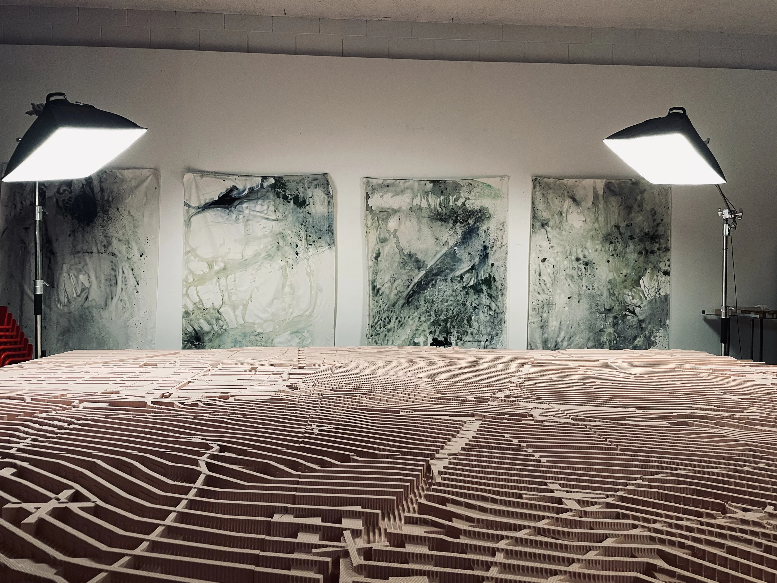 An art studio with three abstract paintings hanging on the wall, large studio lights illuminating, and a detailed cnc sculpture in the foreground.