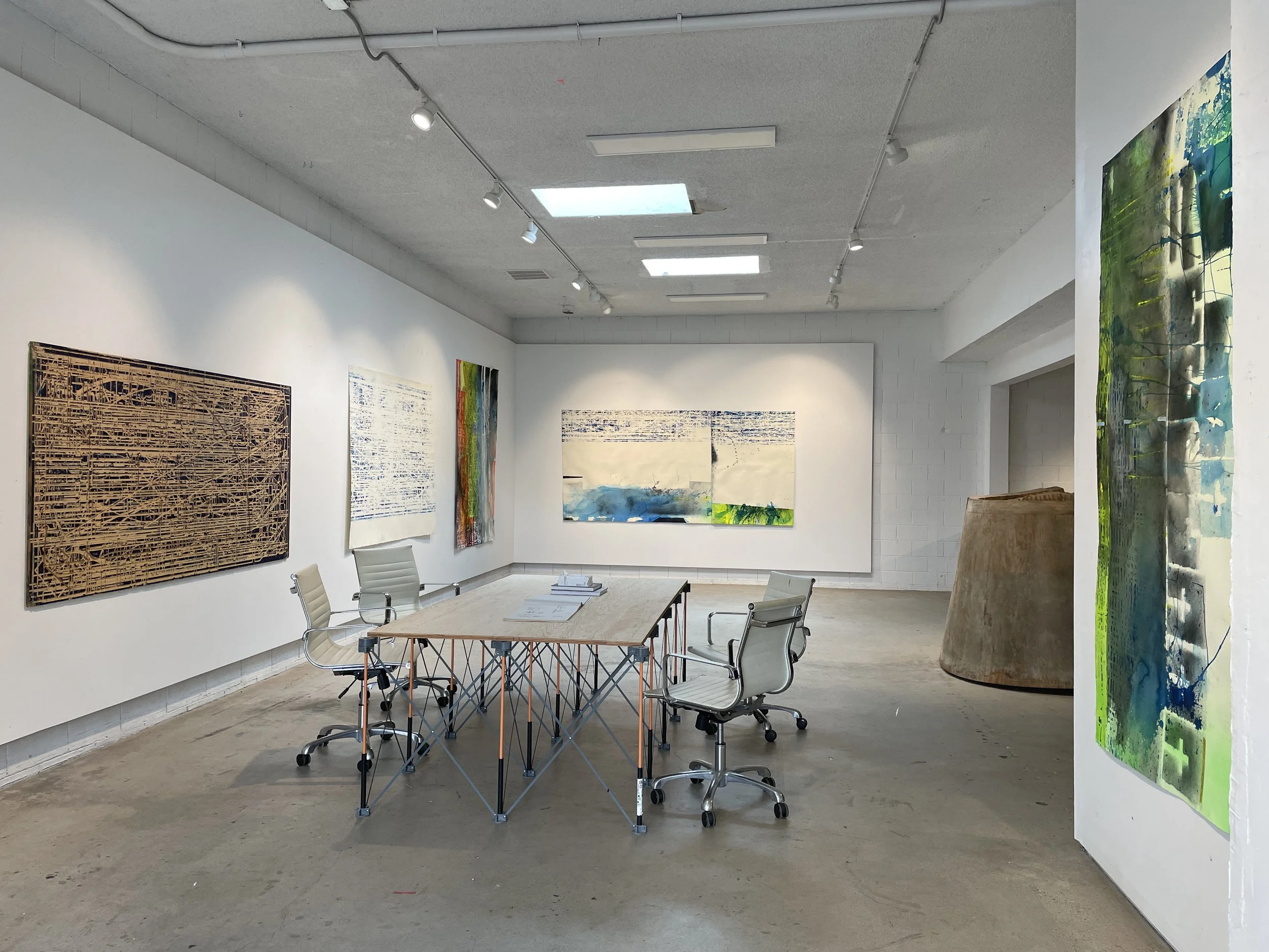 An art gallery with modern abstract paintings hanging on white walls, a wooden table with chairs, and skylights providing natural light.