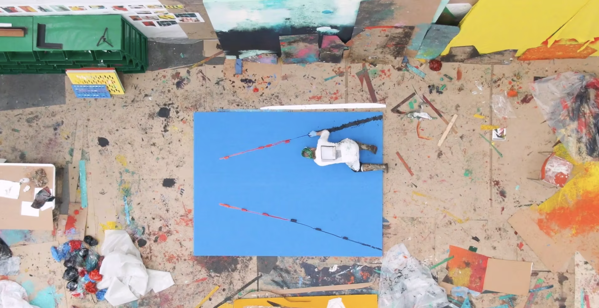 Top-down view of an art studio with a person working on a blue canvas, scattered paint supplies, and colorful abstract paintings in the background.