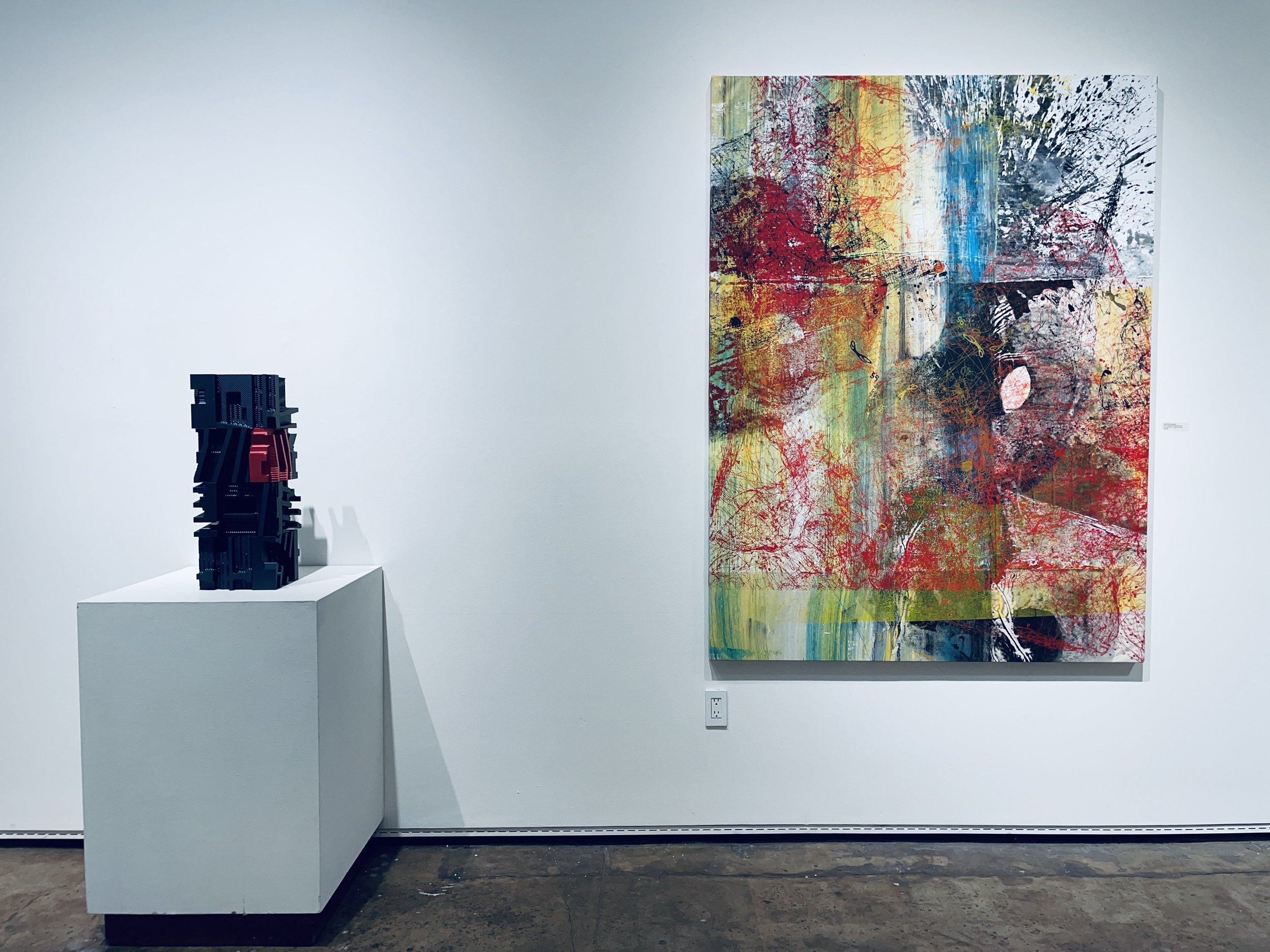 An art gallery features a anodized aluminium sculpture on a white pedestal to the left and a large colorful abstract painting on the white wall to the right.