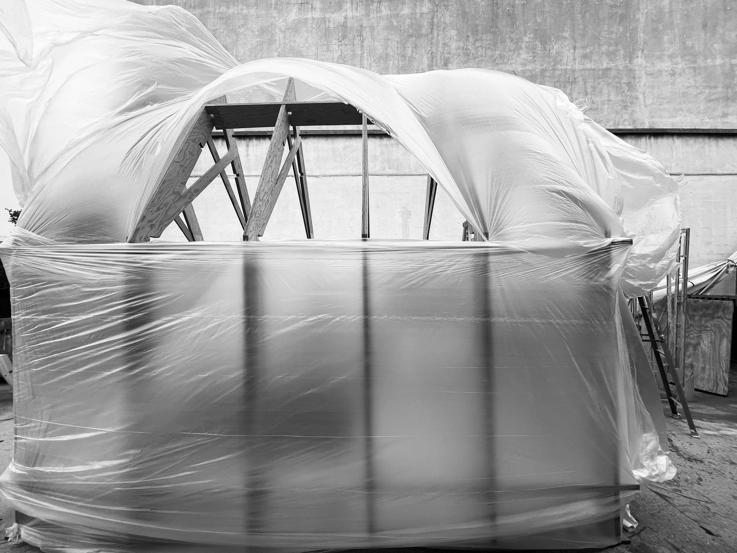Partially built greenhouse structure wrapped in plastic sheeting and supported by wooden and metal scaffolding.