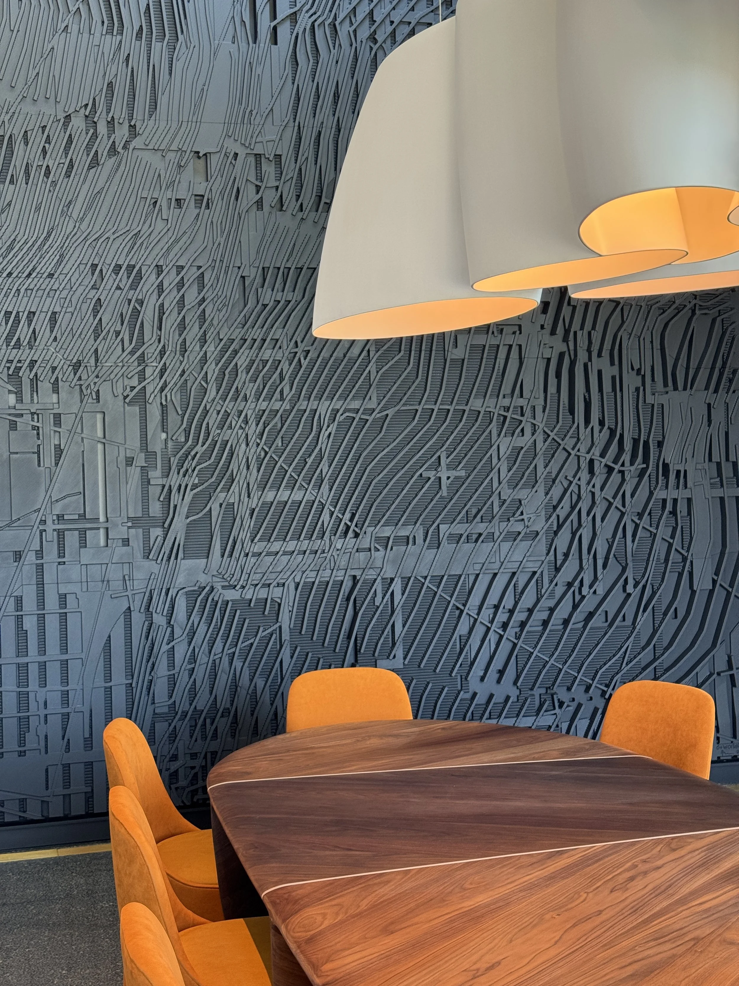 Modern conference room with a wooden table, orange chairs, large pendant lights, and a dark textured wall with abstract patterns.