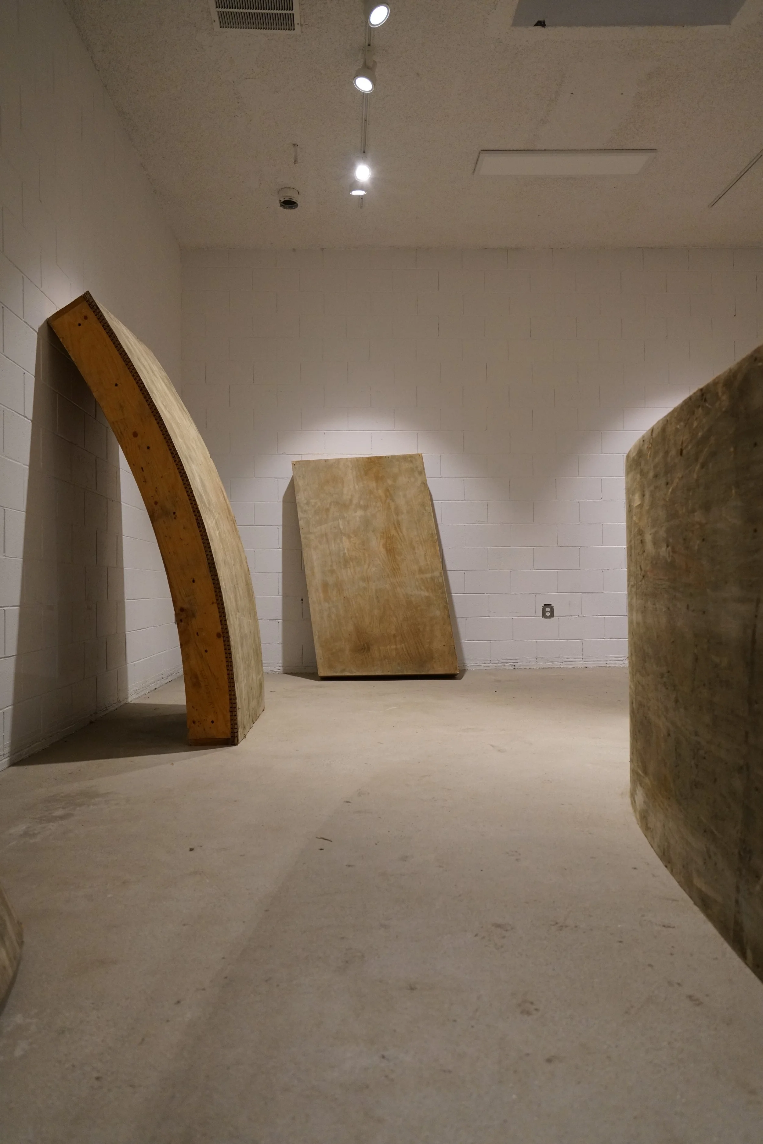 Empty room with white painted brick wall, concrete floor, and three large wooden structures leaning against and near the wall. Overhead lighting is visible.