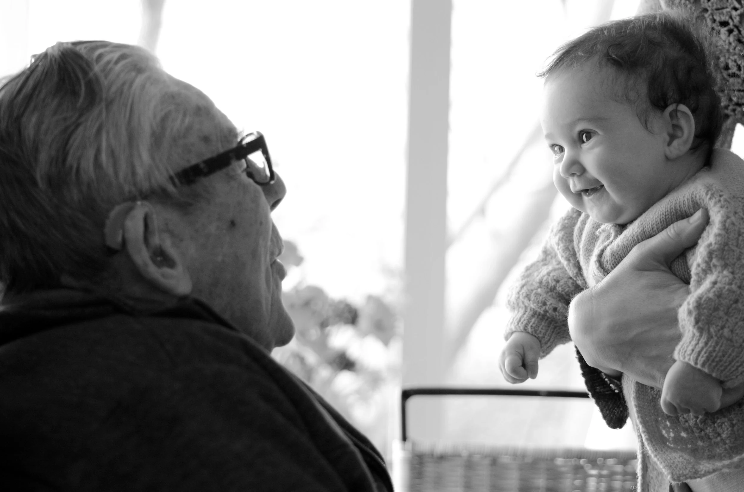 An elderly person wearing glasses looking at a happy young child being held up by an adult.