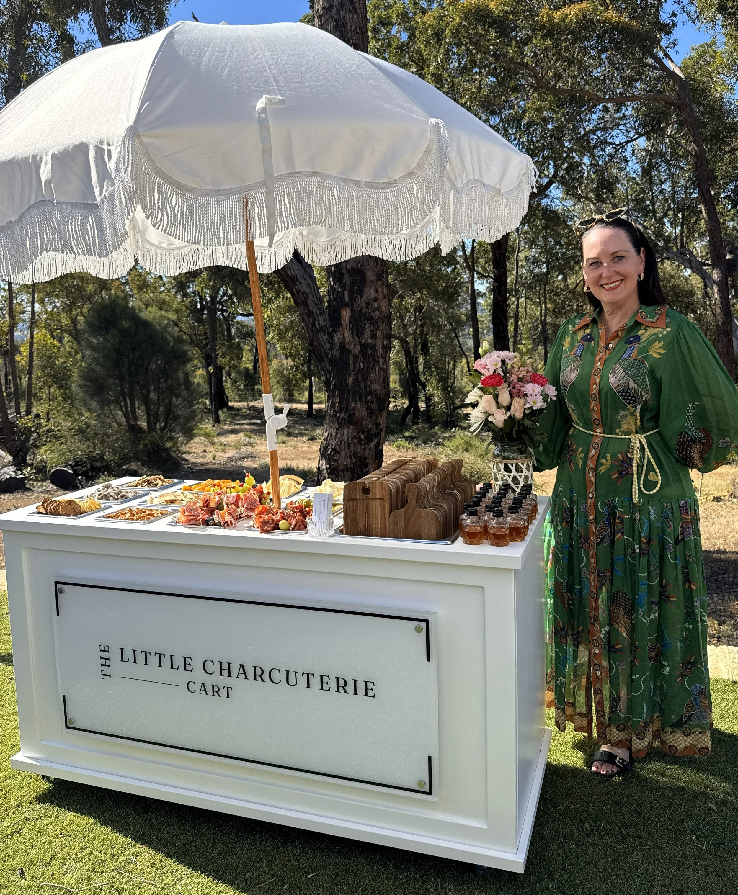 A woman in a colorful dress standing behind a white cart with a sign that reads 'The Little Charcuterie Cart,' outdoor setting with trees, and offering a variety of snacks and drinks under a white parasol.