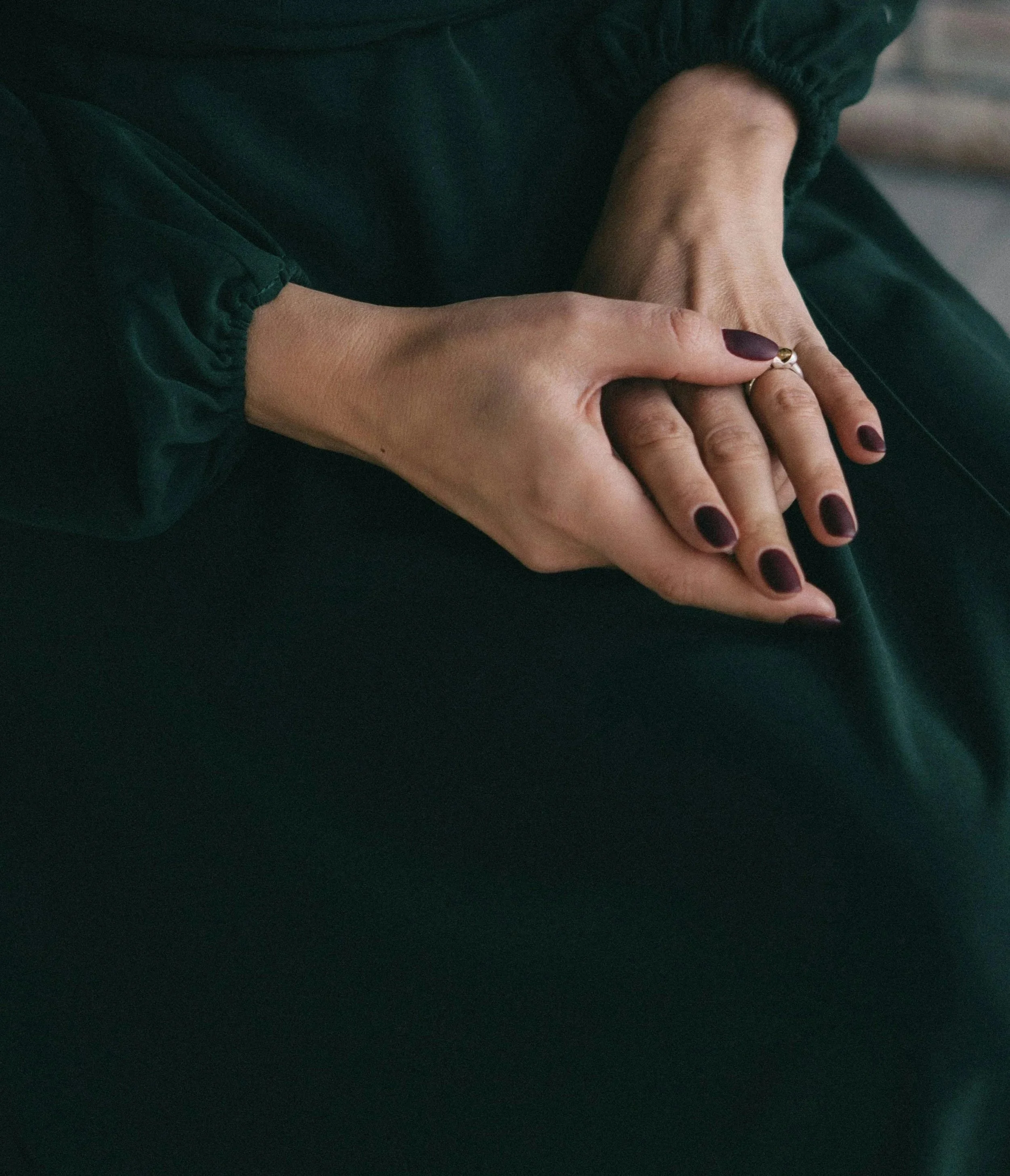 Nahaufnahme von Händen, die auf den Knien ruhen, mit dunklen Nagellack und einem Ring an der rechten Hand.