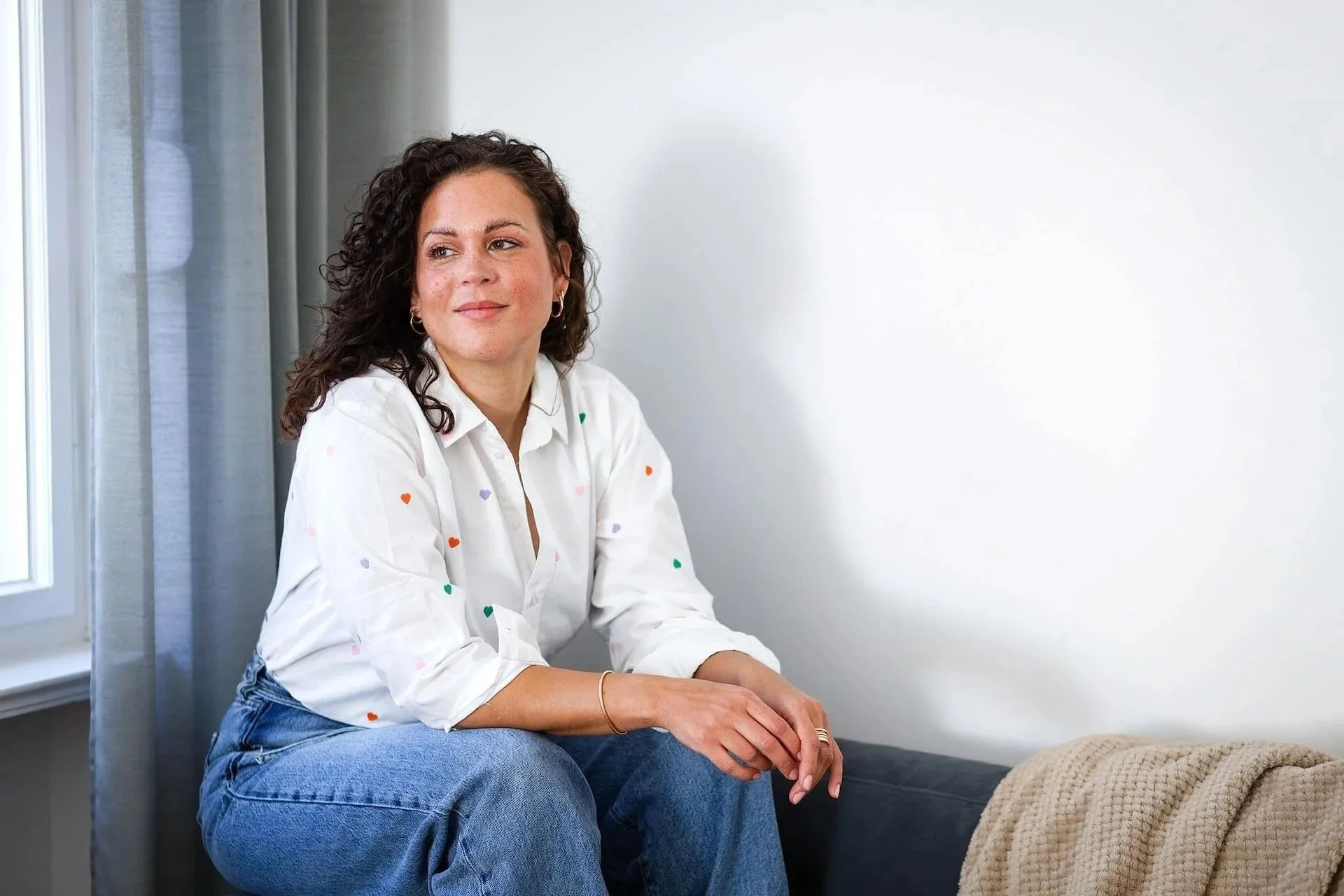 Eine junge Frau mit dunklen, lockigen Haaren sitzt auf einer Couch, trägt ein weißes Hemd mit kleinen bunten Herzen und schaut nach links.