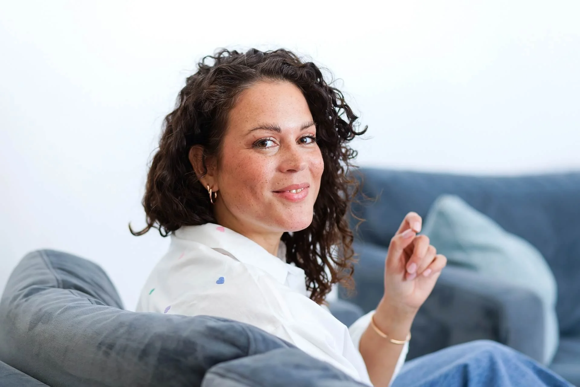 Eine Frau mit lockigem braunem Haar sitzt entspannt auf einem grauen Sofa, trägt eine weiße Bluse mit bunten Herzchen und lächelt freundlich in die Kamera.