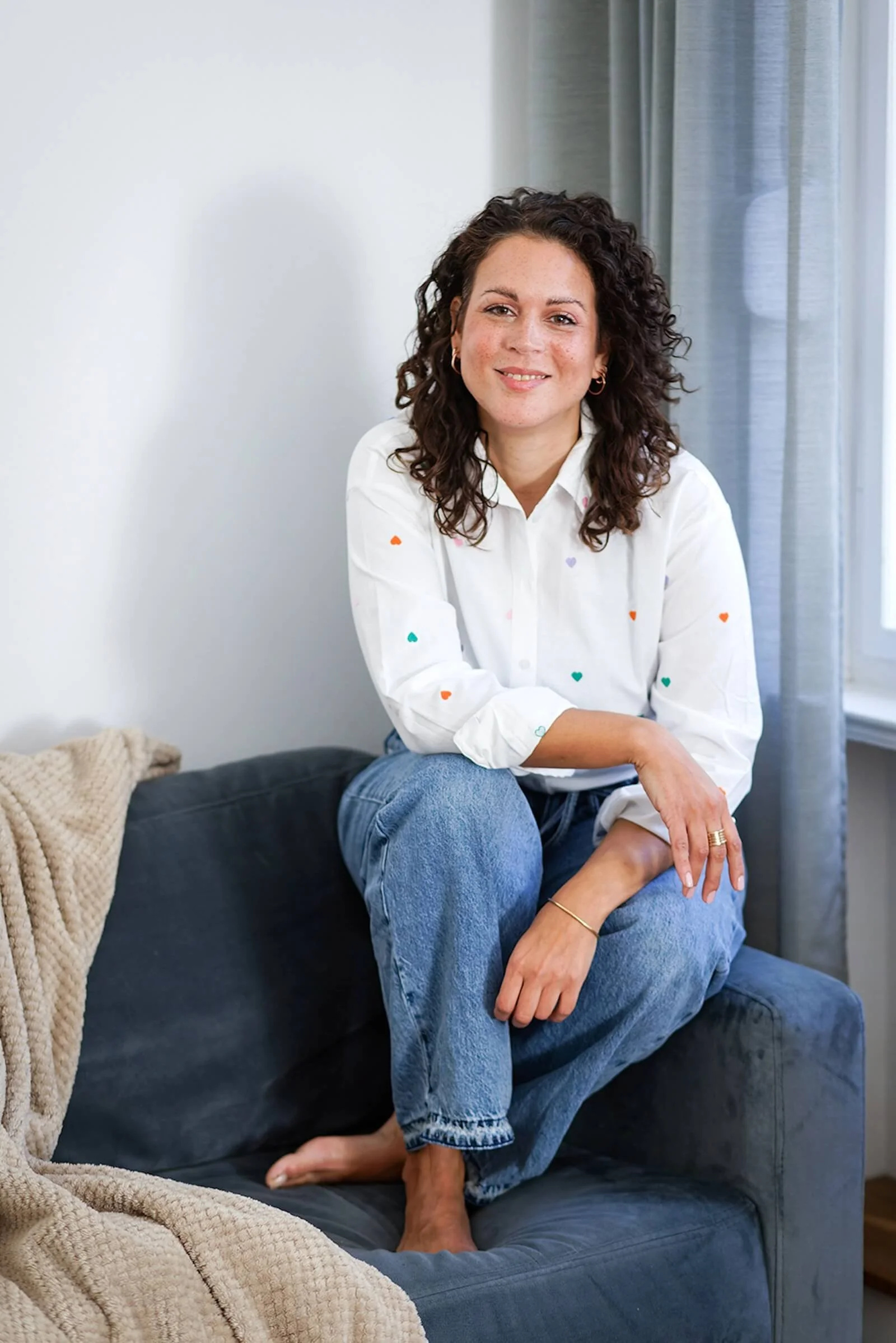 Eine lächelnde Frau mit lockigen braunen Haaren sitzt entspannt auf einem dunklen Sofa, trägt eine weiße Bluse mit kleinen bunten Herzmustern und blaue Jeans, neben einem beige gestrickten Kissen.