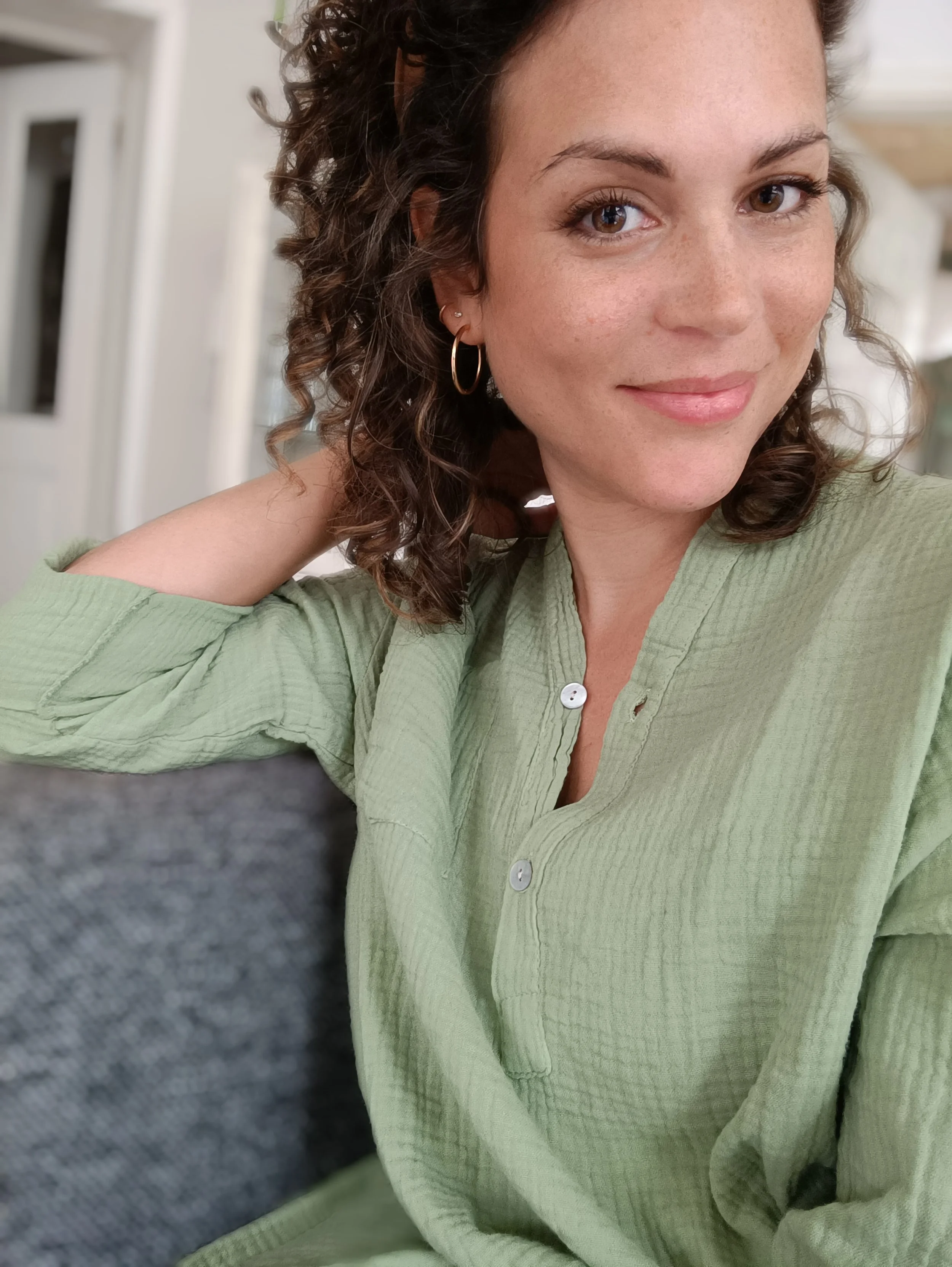 Ein junges Mädchen mit lockigem braunem Haar trägt einen grünen Button-Down-Hemd und goldene Ohrringe, sitzt auf einem Sofa, lächelt leicht und schaut in die Kamera.