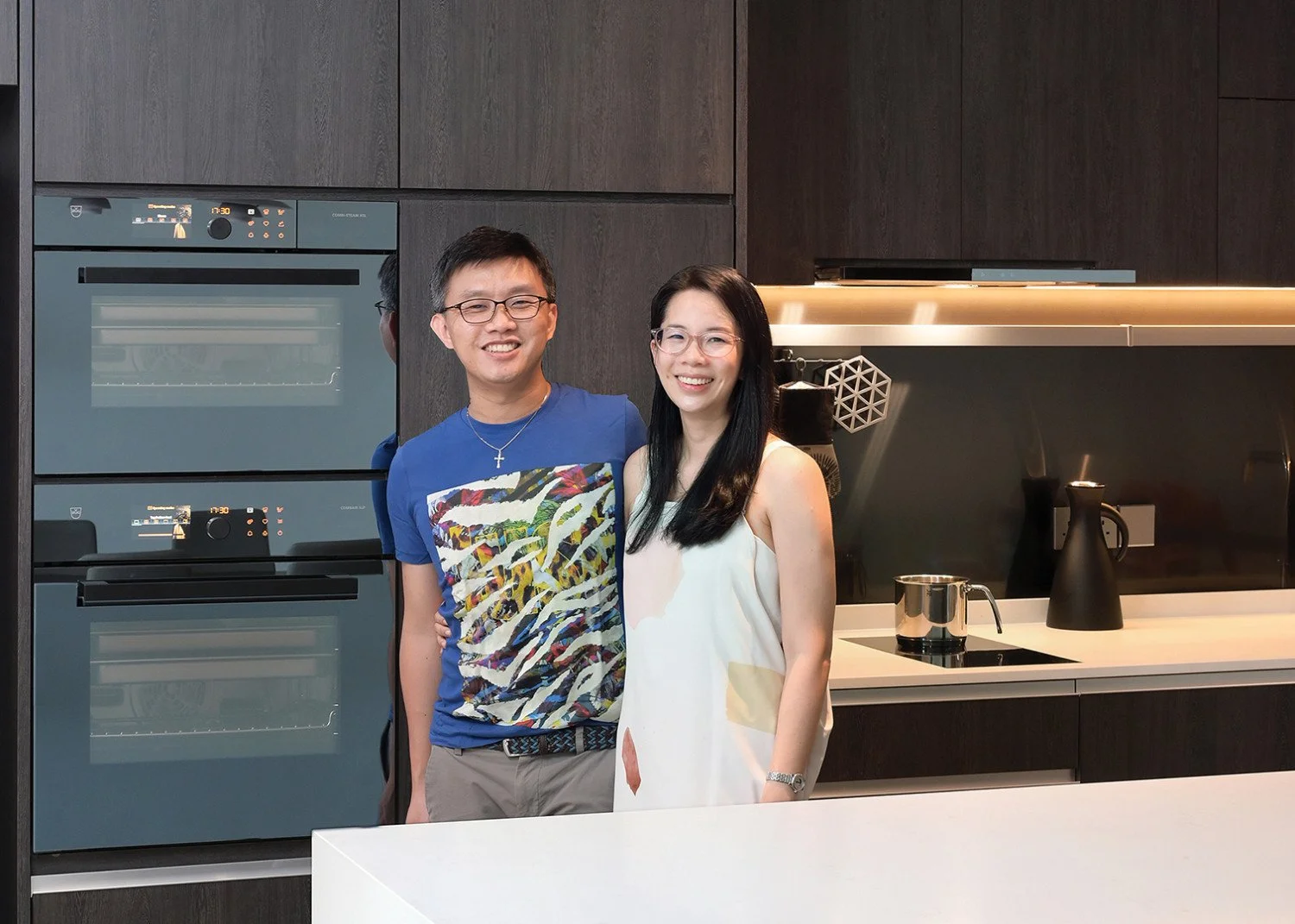A smiling man and woman standing side by side in a modern kitchen with dark wood cabinets, a built-in oven, and a stovetop, posing for a photo.