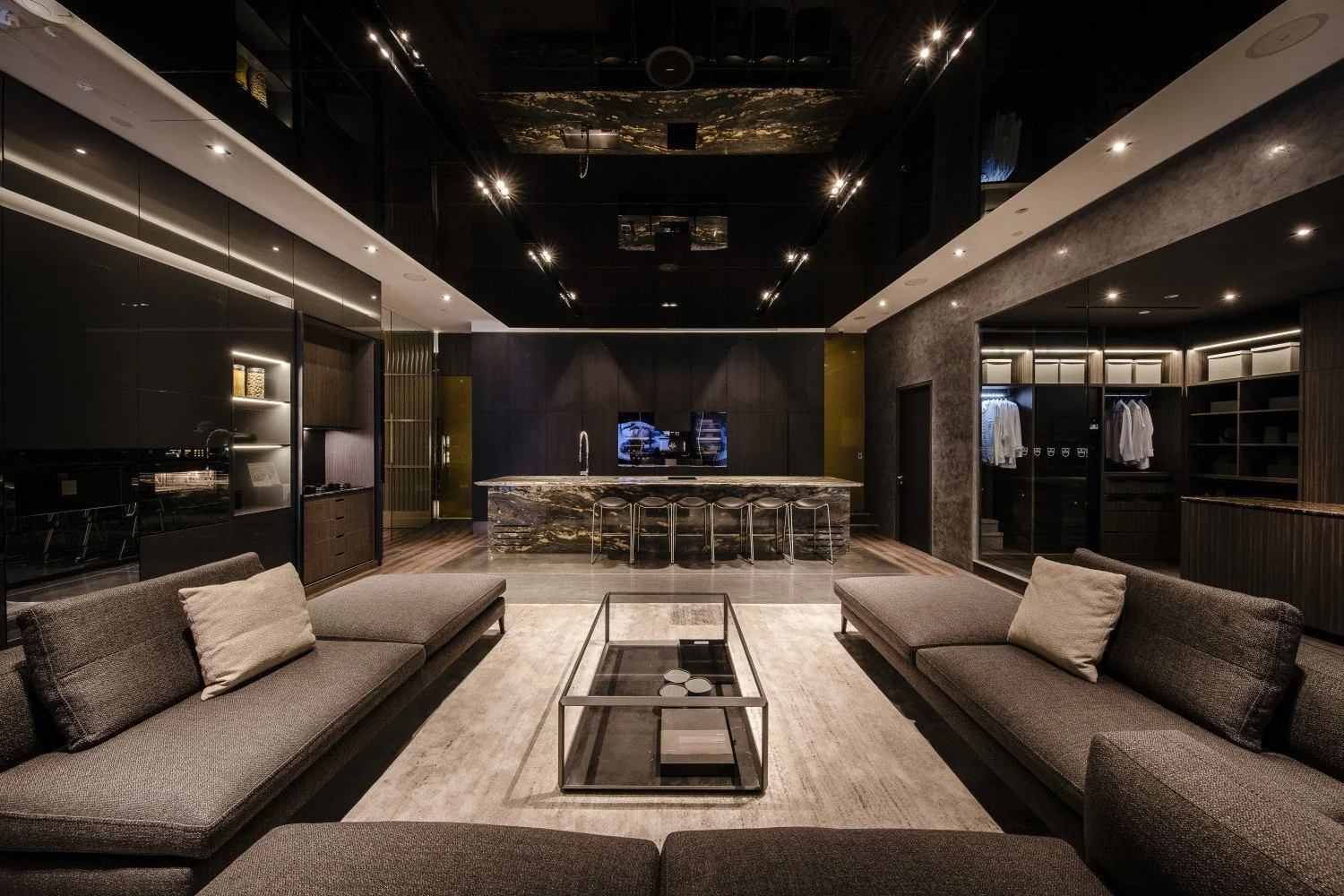 Luxury modern living room with dark walls, a marble kitchen island, gray sofas with cushions, and a minimalist glass coffee table, illuminated by recessed ceiling lights.
