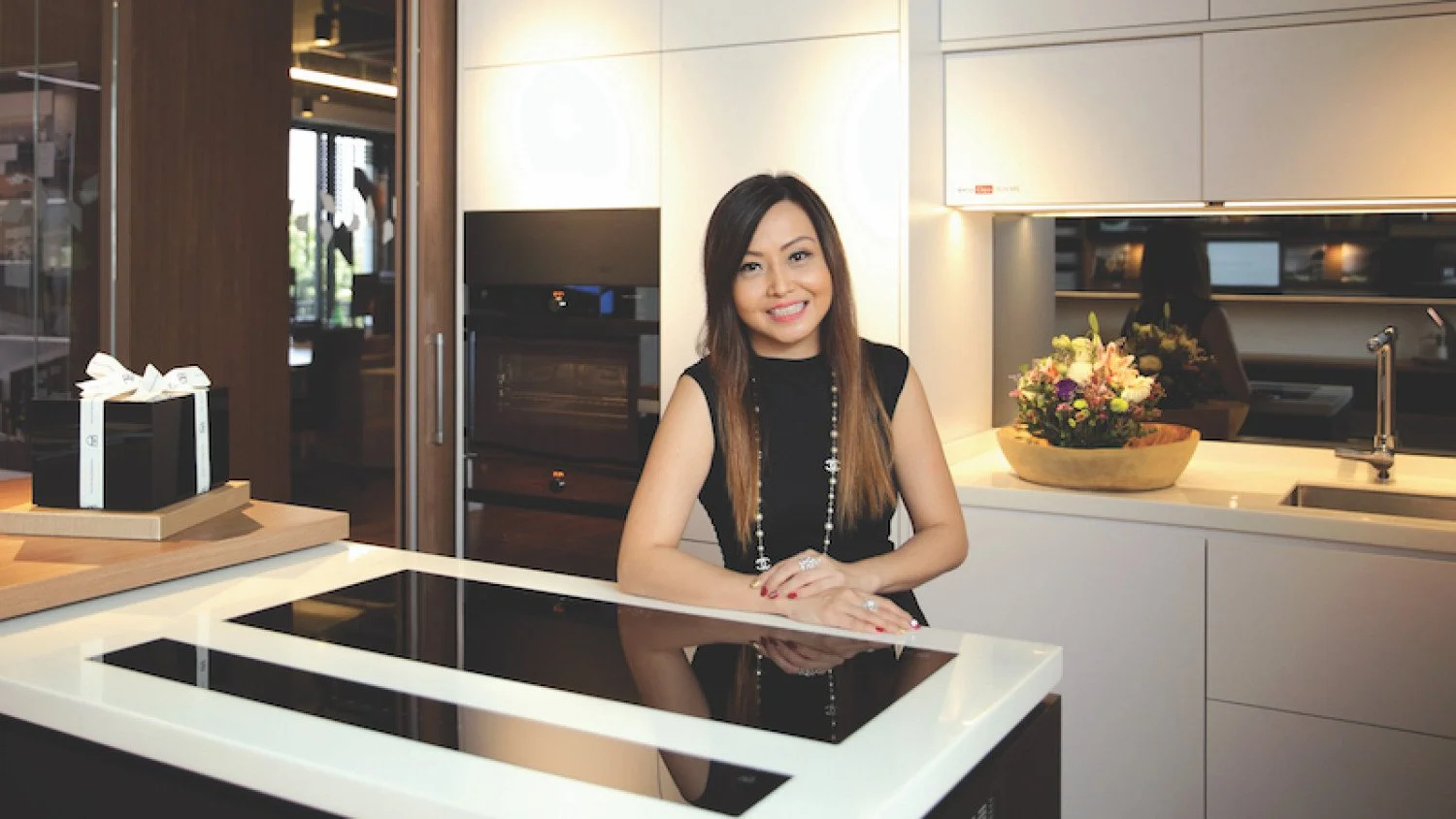 A woman with long brown hair wearing a black sleeveless top and a jewelry necklace, standing in a modern kitchen with white cabinets, a sink, and a flower arrangement on the counter.