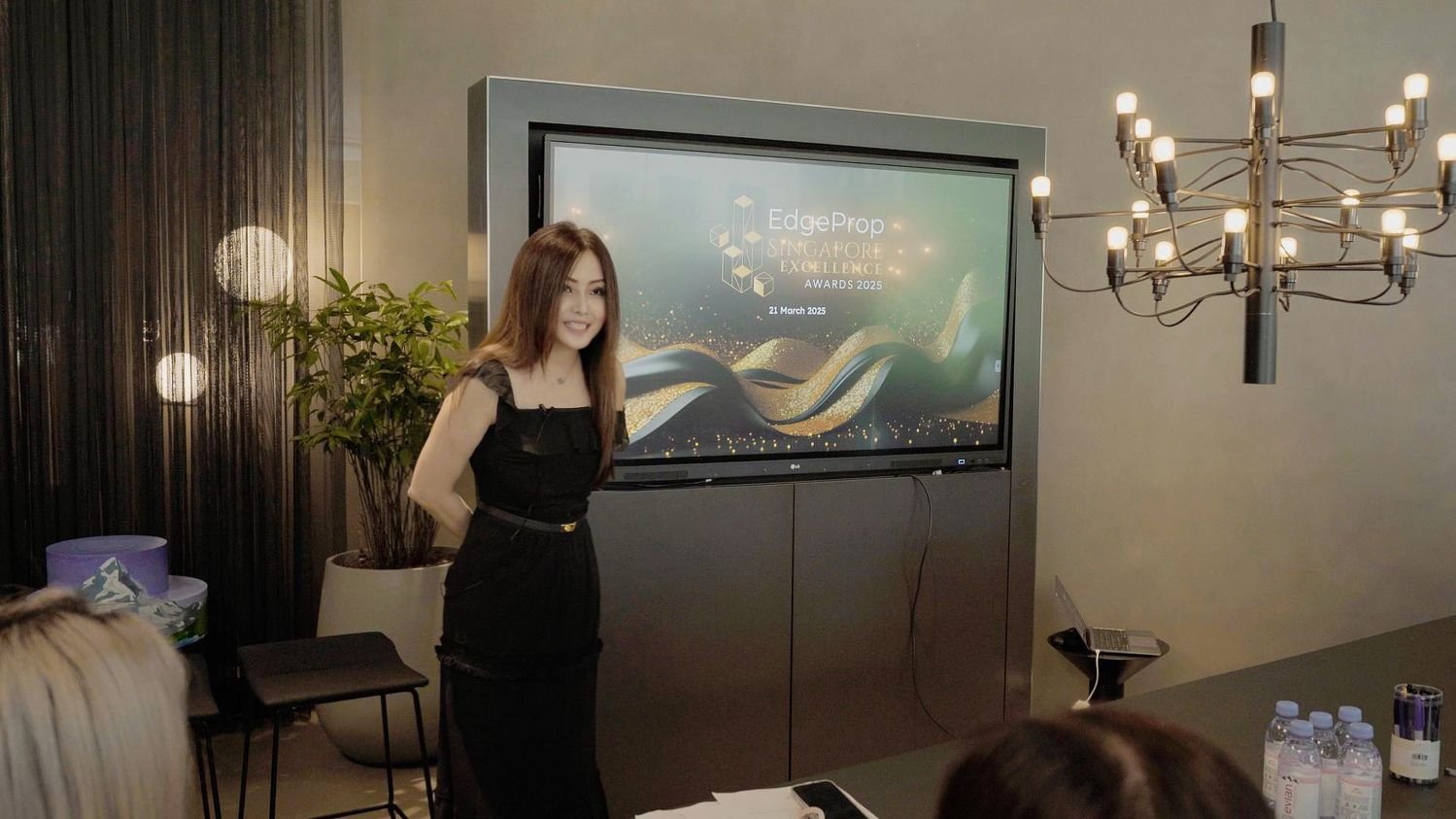 Woman presenting at a conference with a screen displaying 'EdgeProp Singapore Excellence Awards 2025' and the date 21 March 2025. The room has a chandelier, potted plant, and bottles of water on the table.