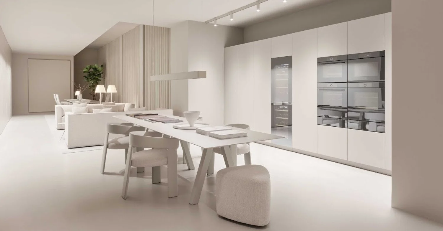 Modern, minimalist open-plan living space with a dining area and kitchen in shades of white and beige. The dining table has chairs and pens, and the kitchen has built-in appliances and cabinets.
