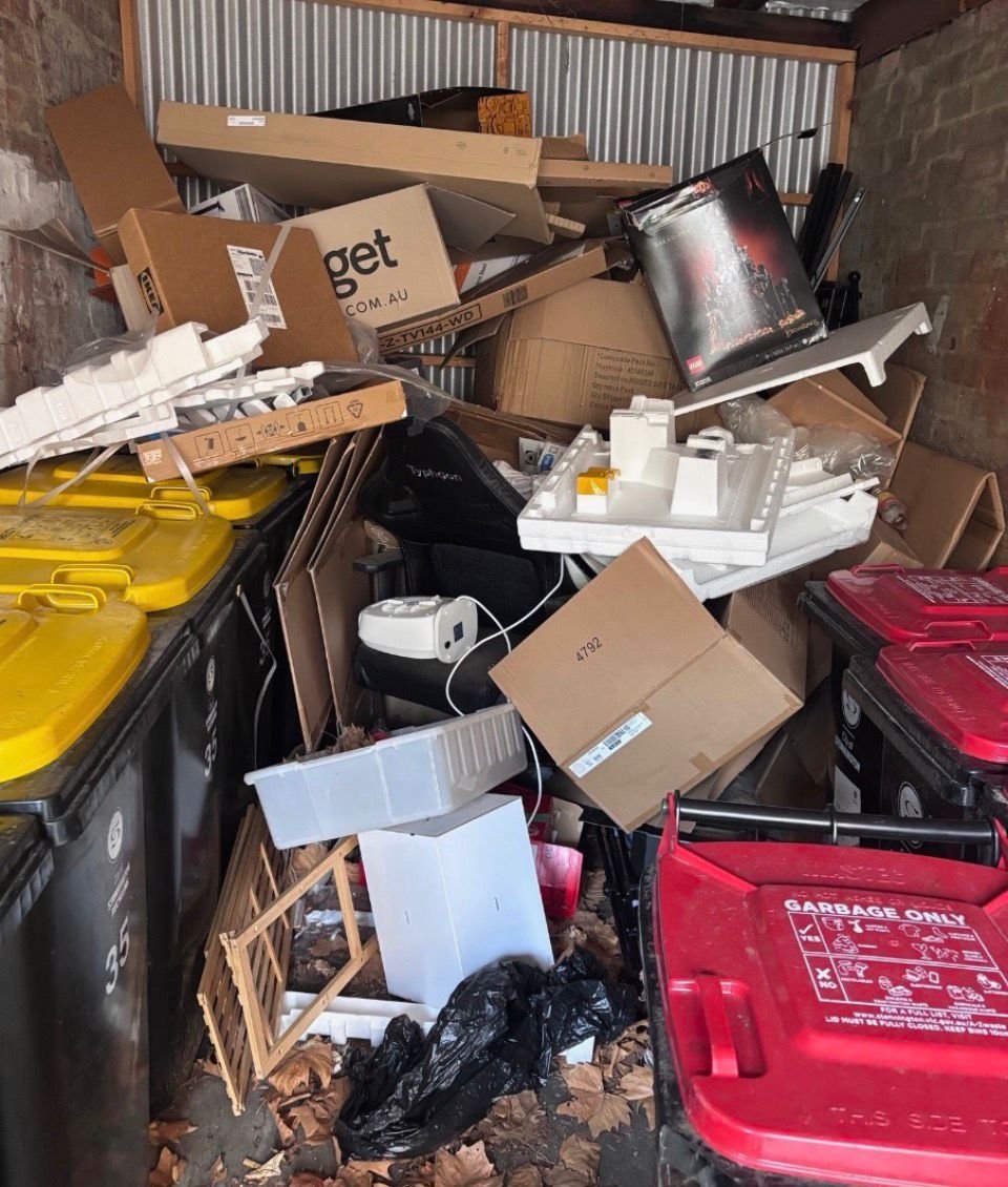 A cluttered storage area with overflowing trash and recycling bins, cardboard boxes, disorganized items, and spilled leaves on the ground.