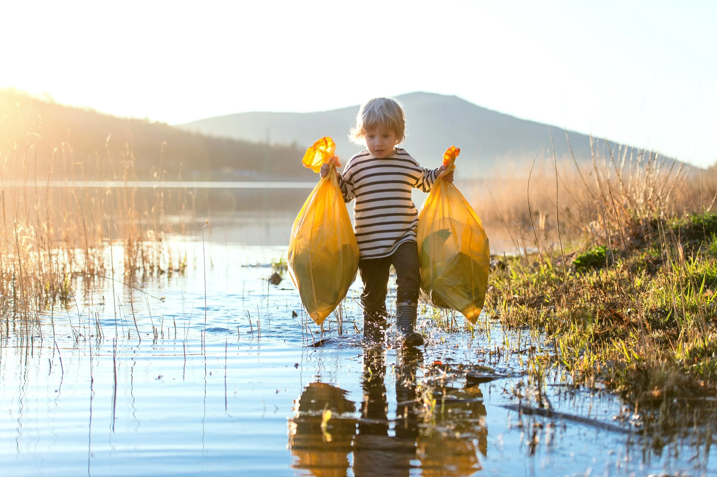 A young child with blonde hair wearing a striped long-sleeve shirt and striped socks wades through shallow water in a marsh or wetland area, holding yellow trash bags in both hands during sunset.