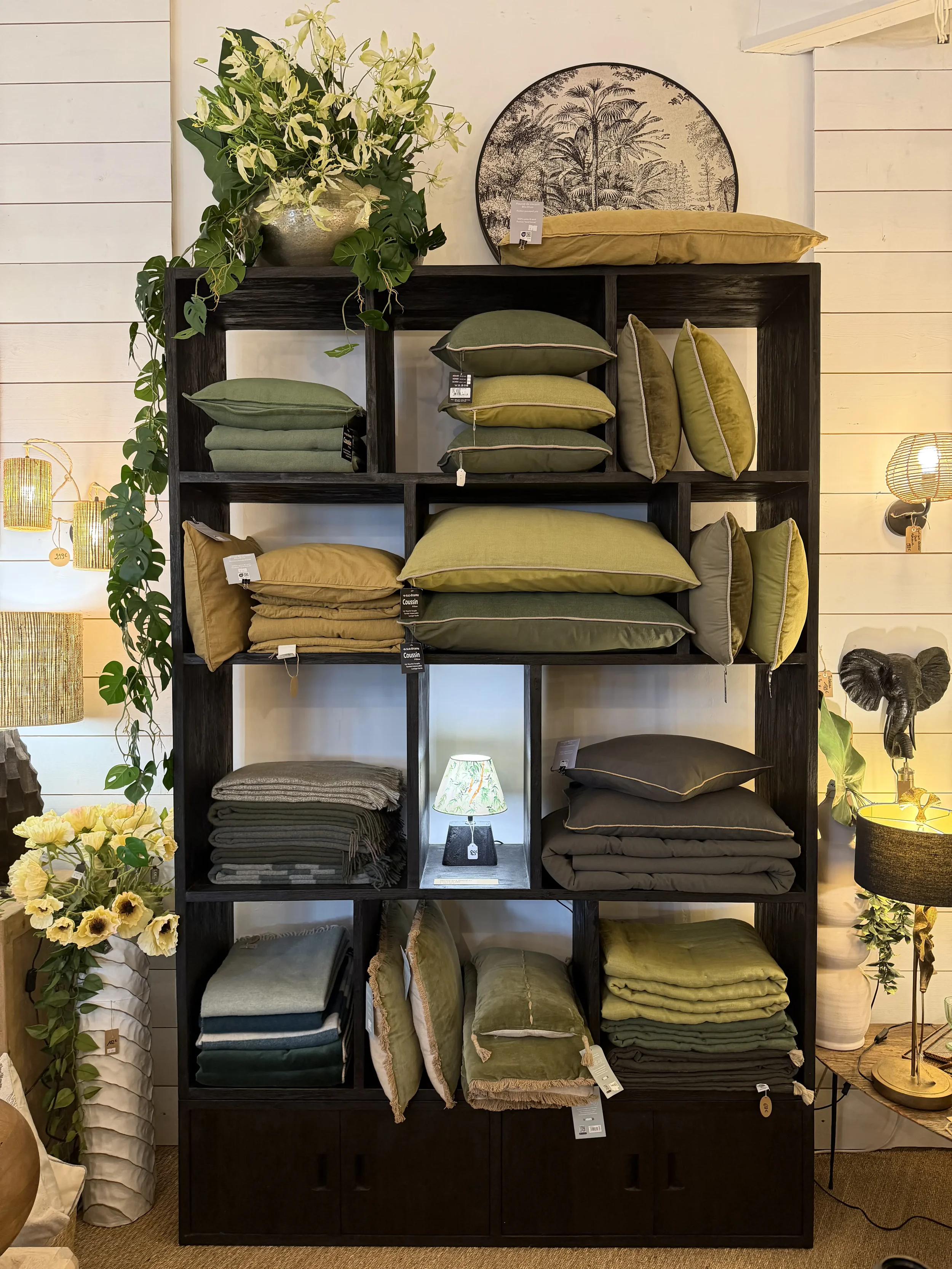 Découvrez une étagère noire remplie d'oreillers et de coussins de différentes tailles et couleurs vert, beige et gris dans une boutique de décoration intérieure. À gauche, une plante verte et une lampe décorative en bas. À droite, une lampe de table et un cadre photographique rond avec un dessin de végétation.