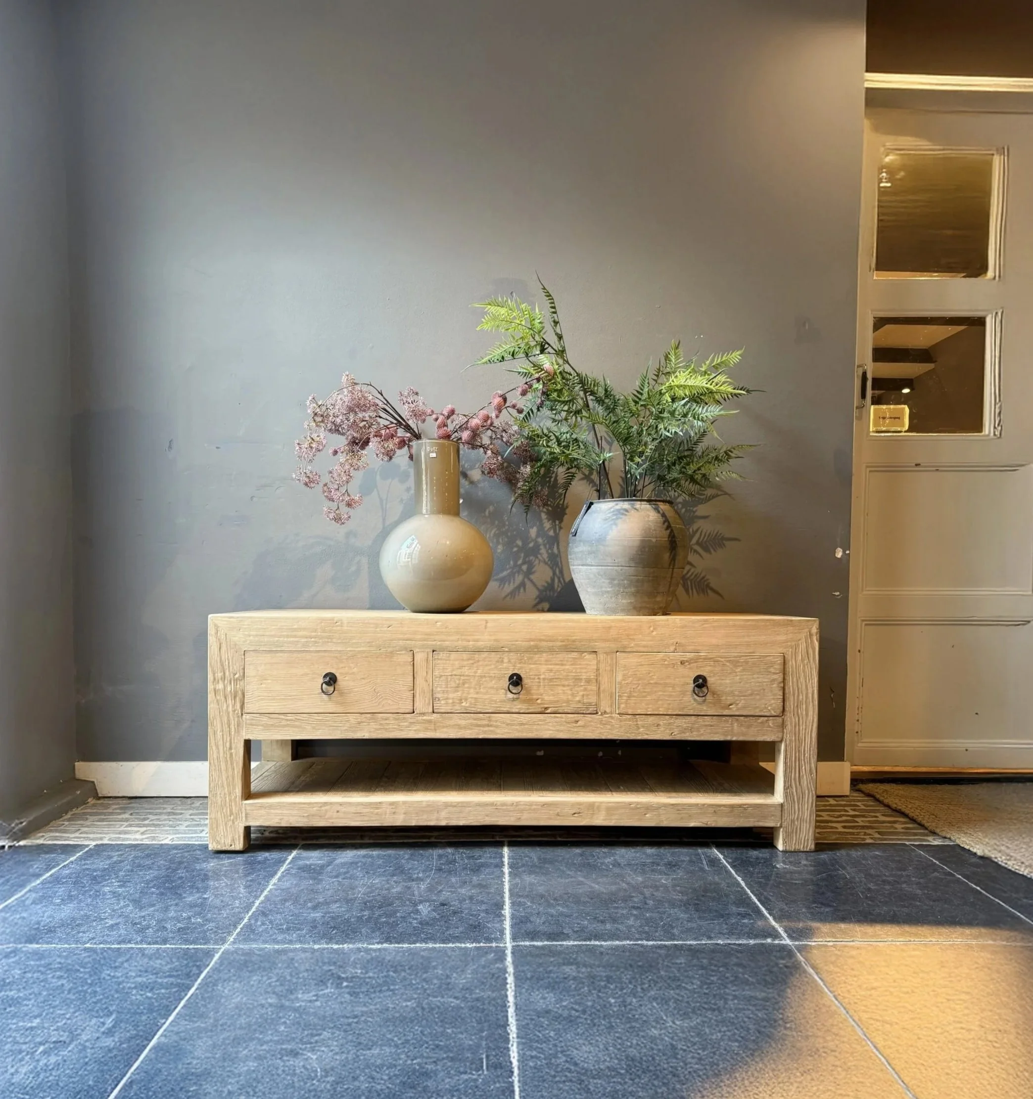 Une console en bois clair avec trois tiroirs, décorée de deux vases avec des plantes et fleurs, sur un sol en carreaux foncés, à côté d'une porte en bois gris.