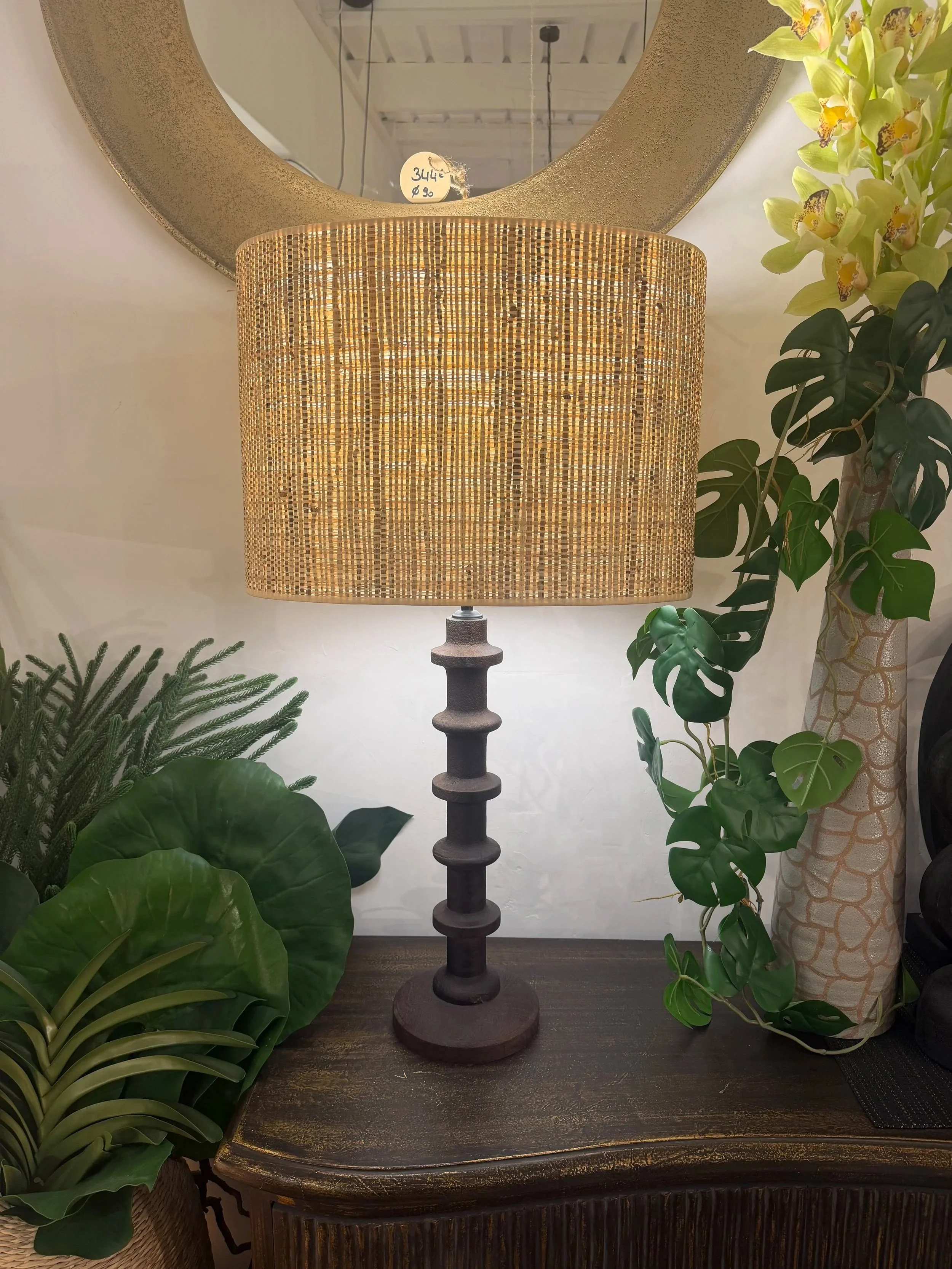 lampe de table avec un pied en bois sculpté et un abat-jour en rotin placé sur une console en bois foncé. Il y a des plantes vertes à gauche et à droite, avec un miroir rond en arrière.