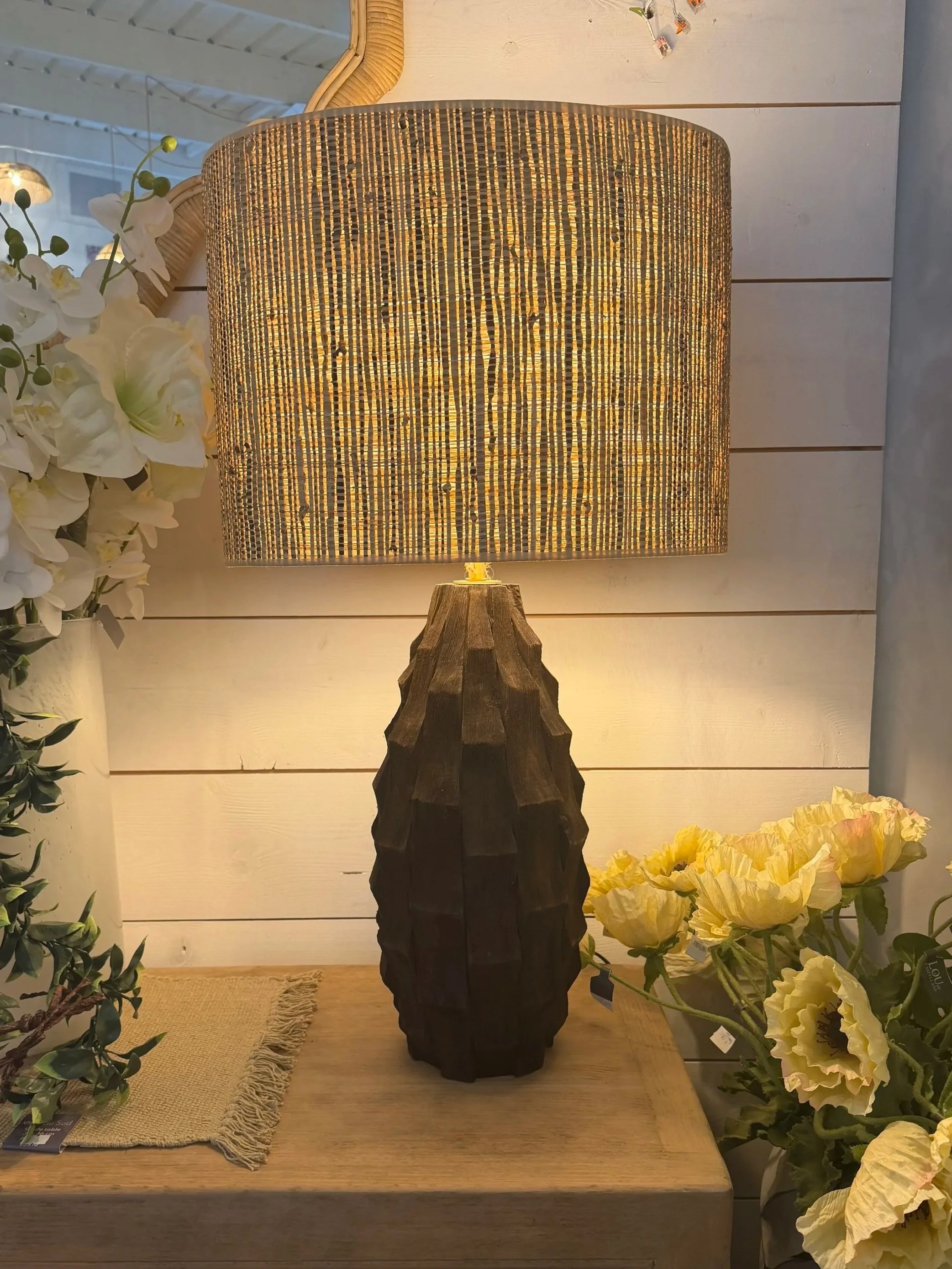 Lampe de table avec base en bois sculpté, abat-jour en tissu lumineux, décorée de faïence et de fleurs artificielles, dans un intérieur décoratif.