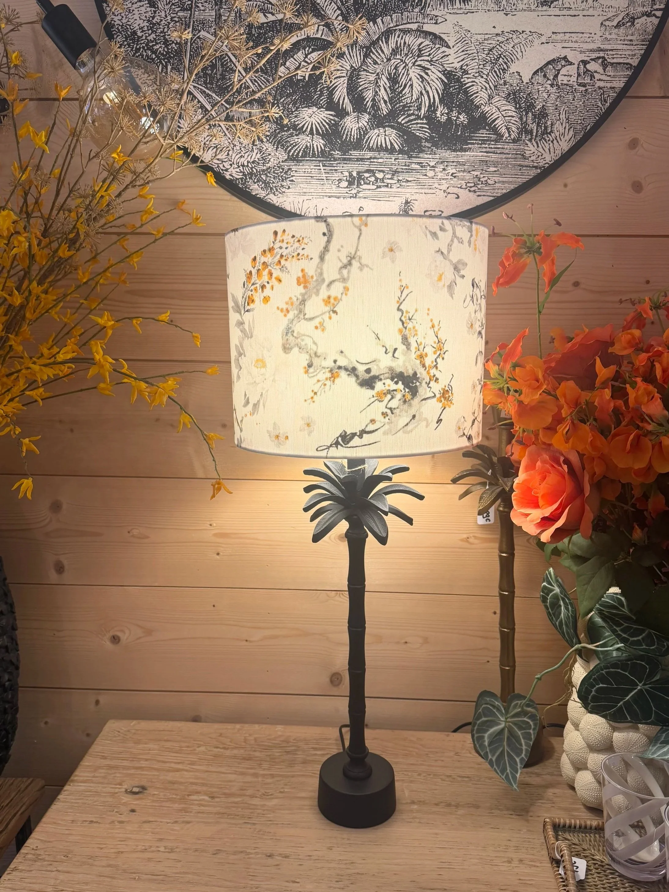 Lampe de table avec un abat-jour blanc à motifs floraux, placée sur une table en bois, entourée de fleurs oranges et vertes.