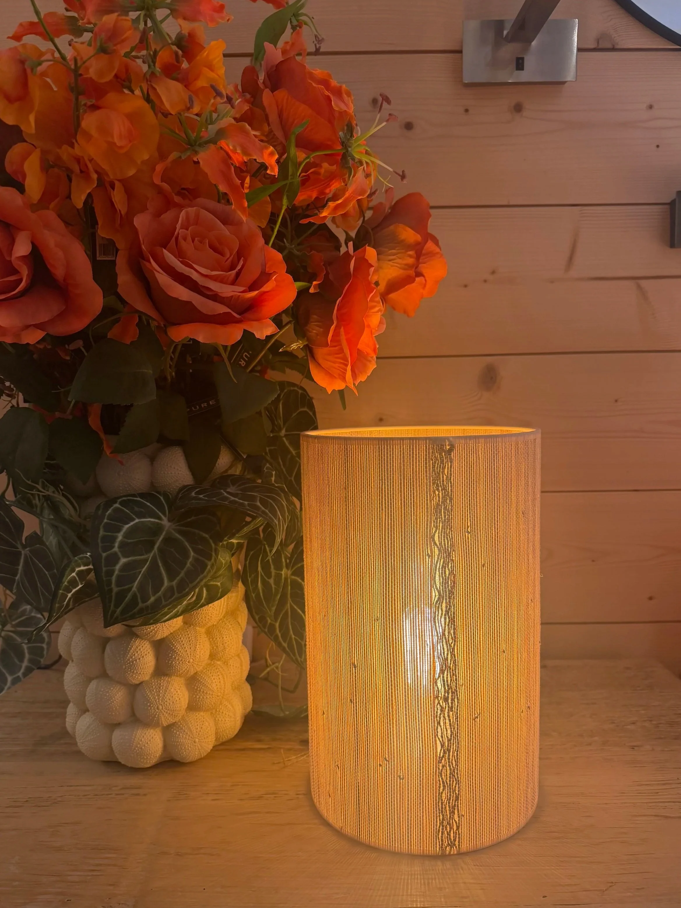 Une lampe cylindrique en tissu éclairée, à côté d'un arrangement de fleurs comprenant des roses orange et d'autres fleurs, sur une surface en bois, avec un mur en bois en arrière-plan.