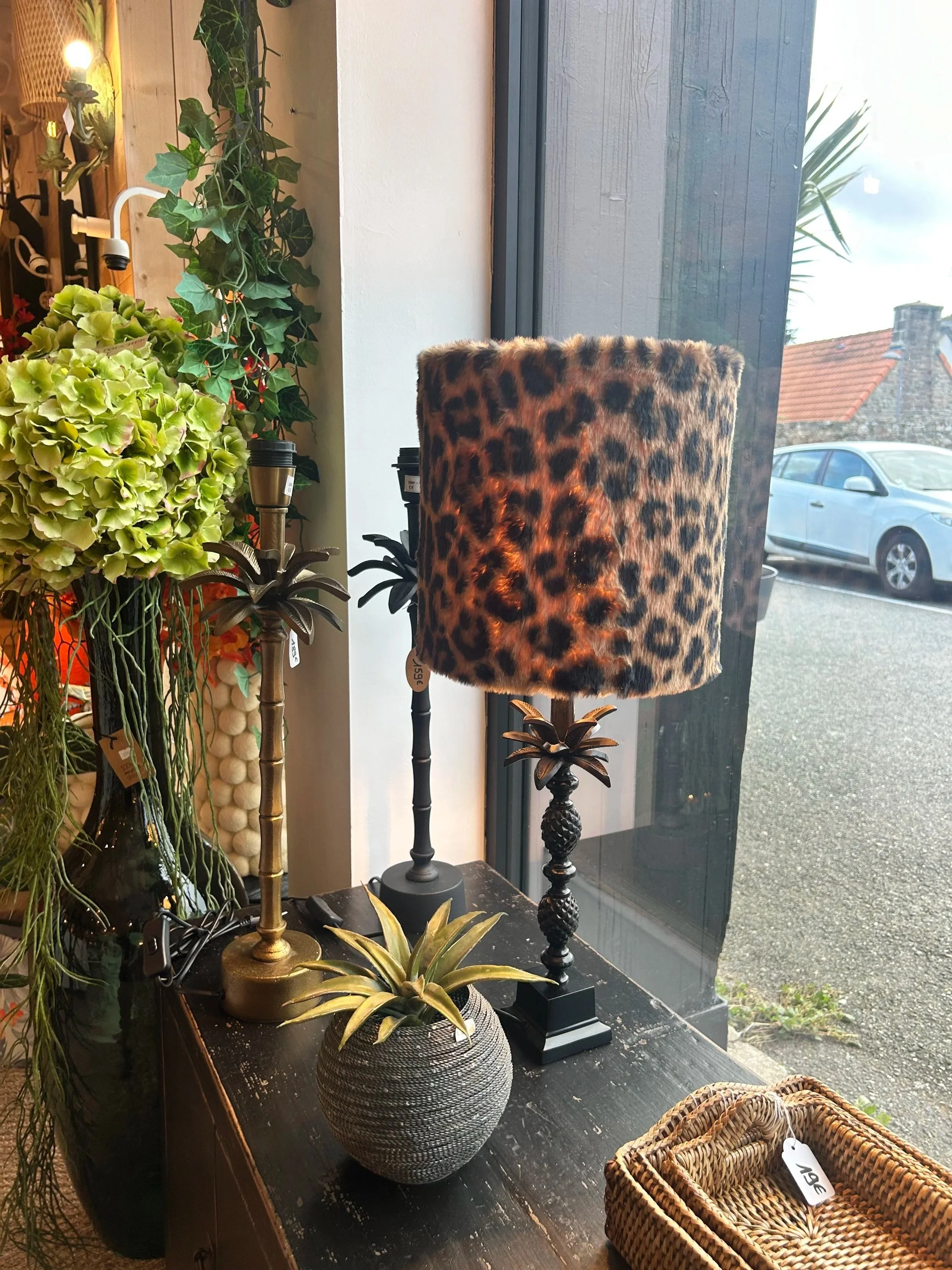 Deux lampes de table, une avec un abat-jour en fausse fourrure à motif léopard, une autre à base de plante en bois, placées sur une surface en bois sombre à côté d'un vase avec une plante verte. Il y a aussi une boîte en rotin avec une étiquette et u