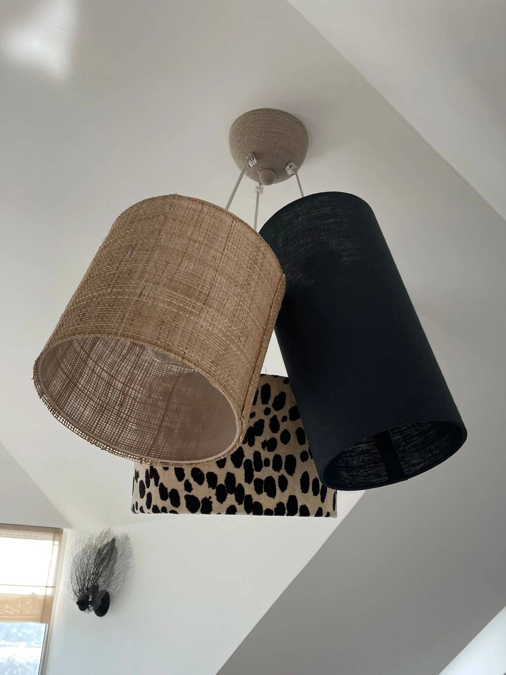Plafonnier avec trois abat-jour de formes et couleurs différentes : un en rotin beige, un en tissu noir, et un en tissu avec motif léopard beige et noir, suspendus sous un plafond blanc.