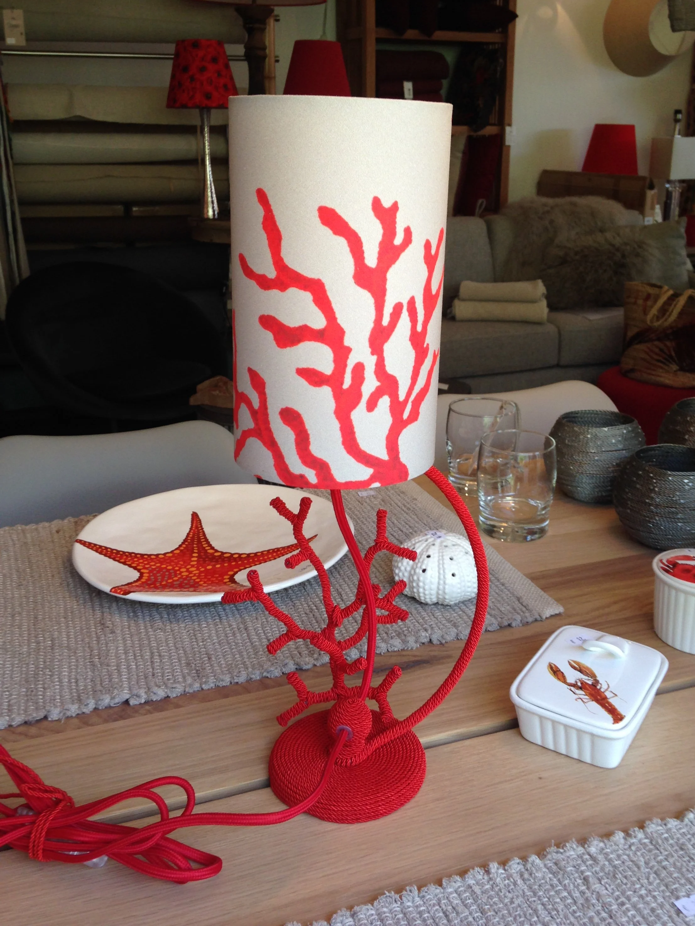 Lampe de table avec abat-jour blanc orné de branches de corail rouges, sur une table en bois dans un magasin d'ameublement.