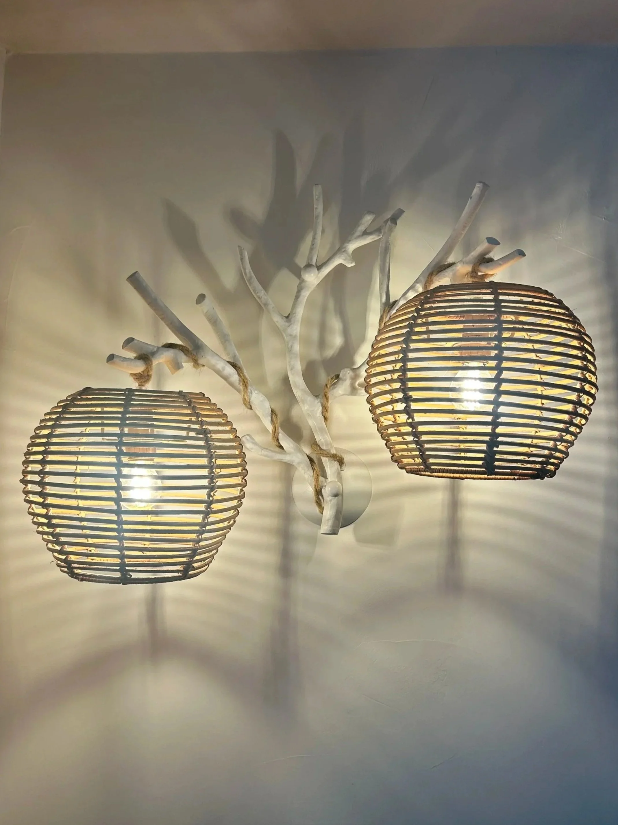 Lampe de plafond avec deux abat-jours en rotin, fixée à une branche décorative en bois peint en blanc qui ressemble à un arbre, créant un jeu d'ombres et de lumières sur le mur.