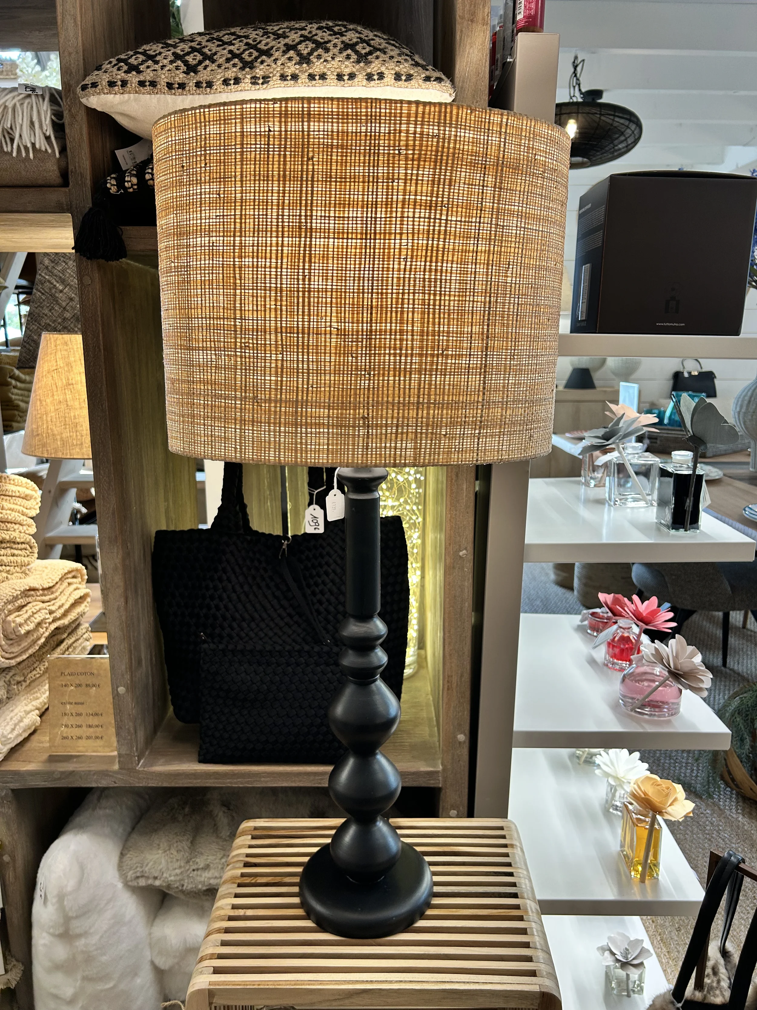 Lampe de table avec un socle noir et un abat-jour en rotin, placée sur une table en bois clair.