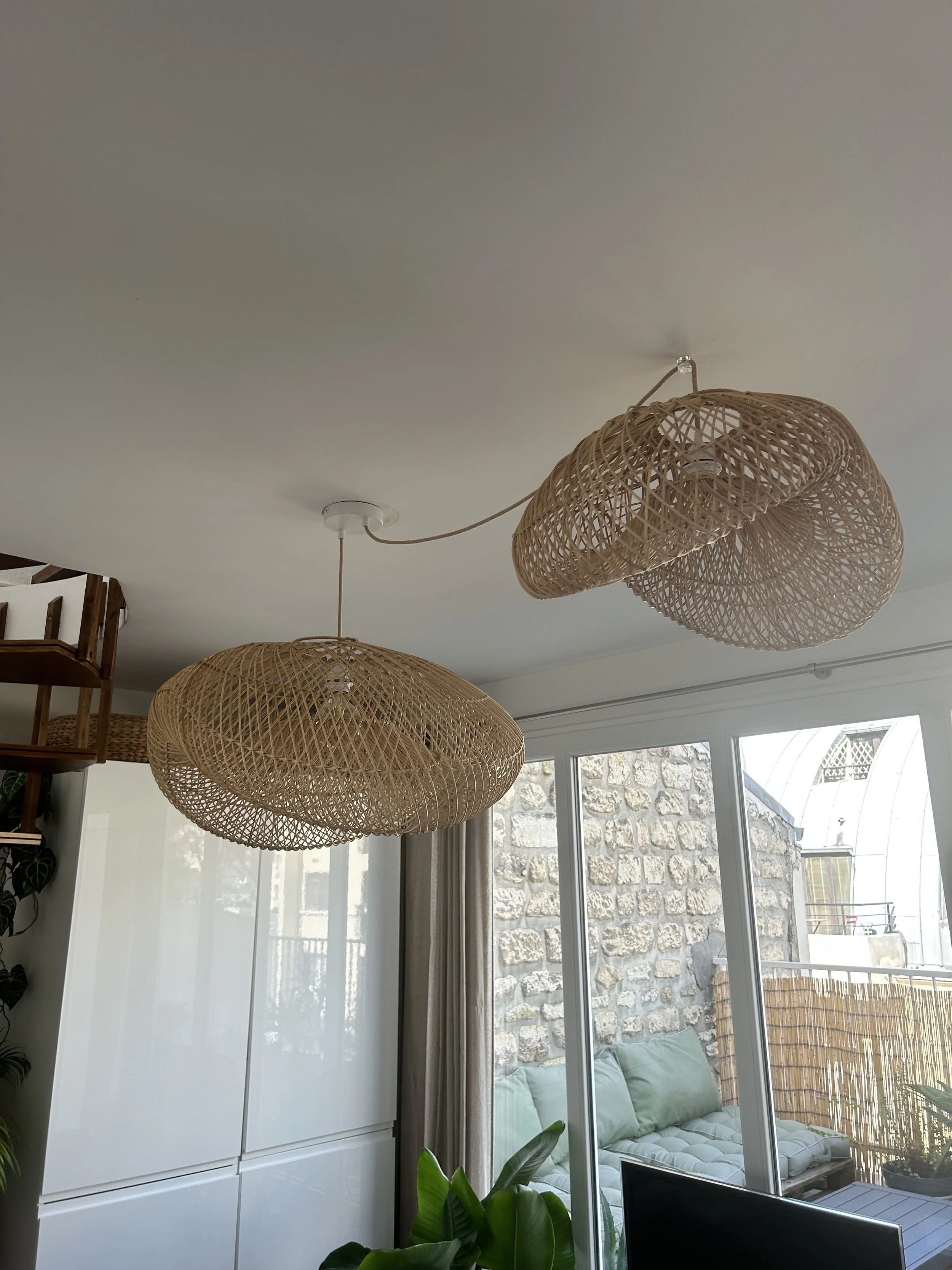 Deux plafonniers en rotin suspendus dans un salon lumineux avec une grande baie vitrée laissant entrer la lumière naturelle.