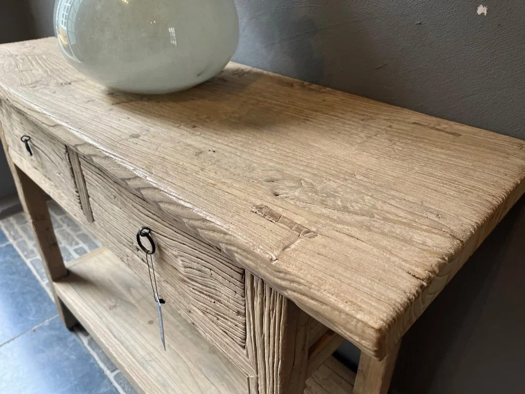 Un meuble en bois brut avec deux tiroirs et une grande surface plate sur le dessus, décoré avec un vase en céramique blanc.