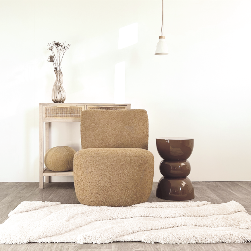 Un coin salon minimaliste avec un fauteuil beige, une petite table en forme d'anneau noir brillant, une console en bois clair avec un vase de fleurs séchées, un tapis blanc, et un luminaire suspendu.