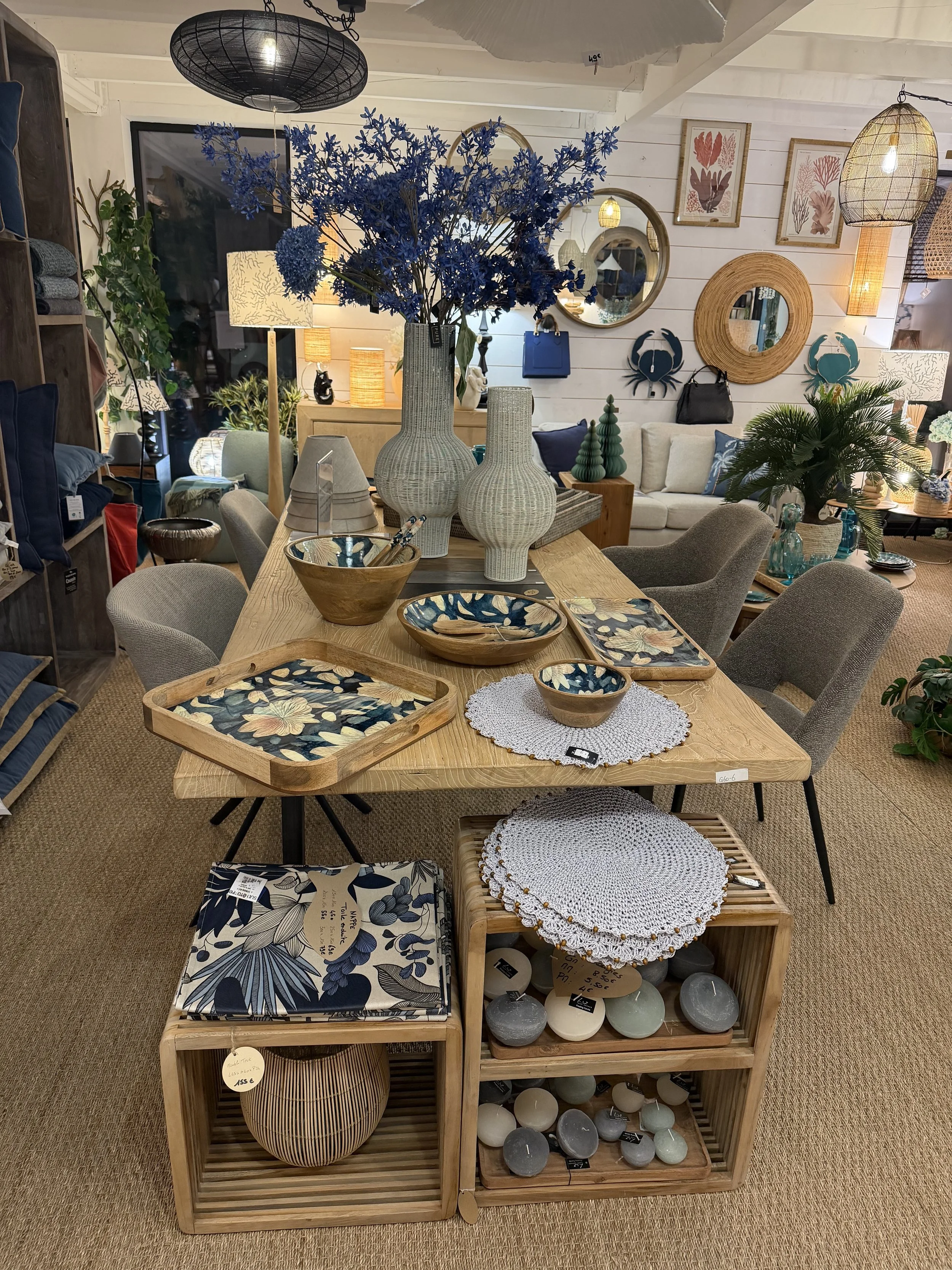 Une table en bois avec des vases, des bols, des plateaux et des sets de table en rotin, tous décorés de motifs floraux ou naturels, dans une boutique d'ameublement et de décoration.