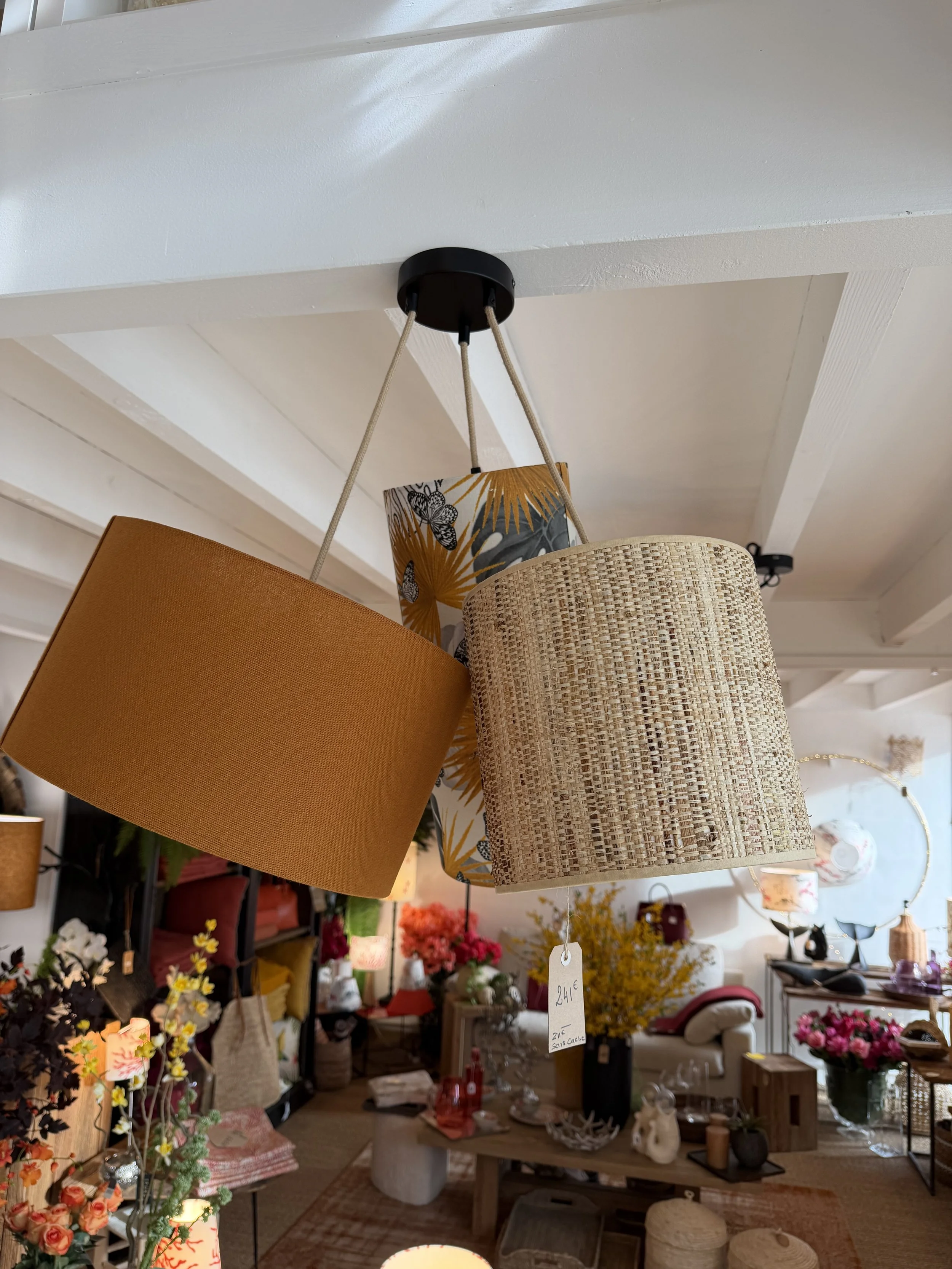 Lustre suspendu avec trois abat-jours de styles différents, dont un en tissu de couleur marron, un en tissu à motifs végétaux et animaux, et un en tissu tissé beige, dans un magasin d'ameublement ou décoration.
