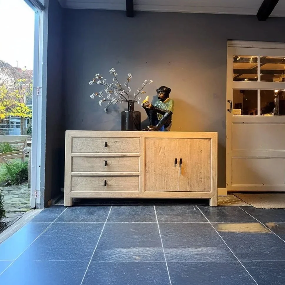 Une commode en bois clair avec une décoration, un vase noir avec des branches de fleurs blanches, une sculpture d'un singe tenant une banane dans un intérieur moderne.