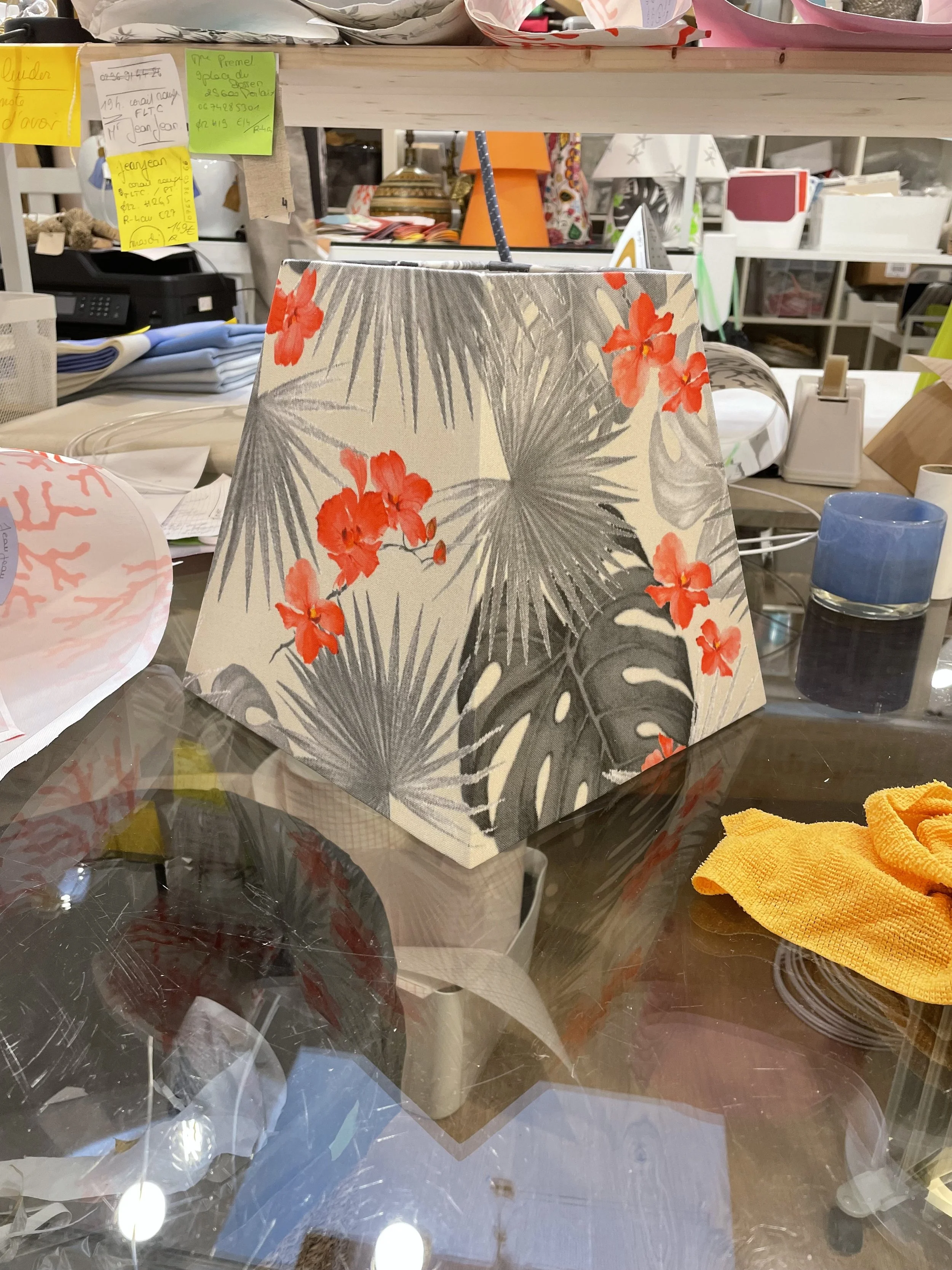Lampadaire de décoration avec motif de feuilles de palmier et fleurs rouges, placé sur une table en verre dans un magasin.