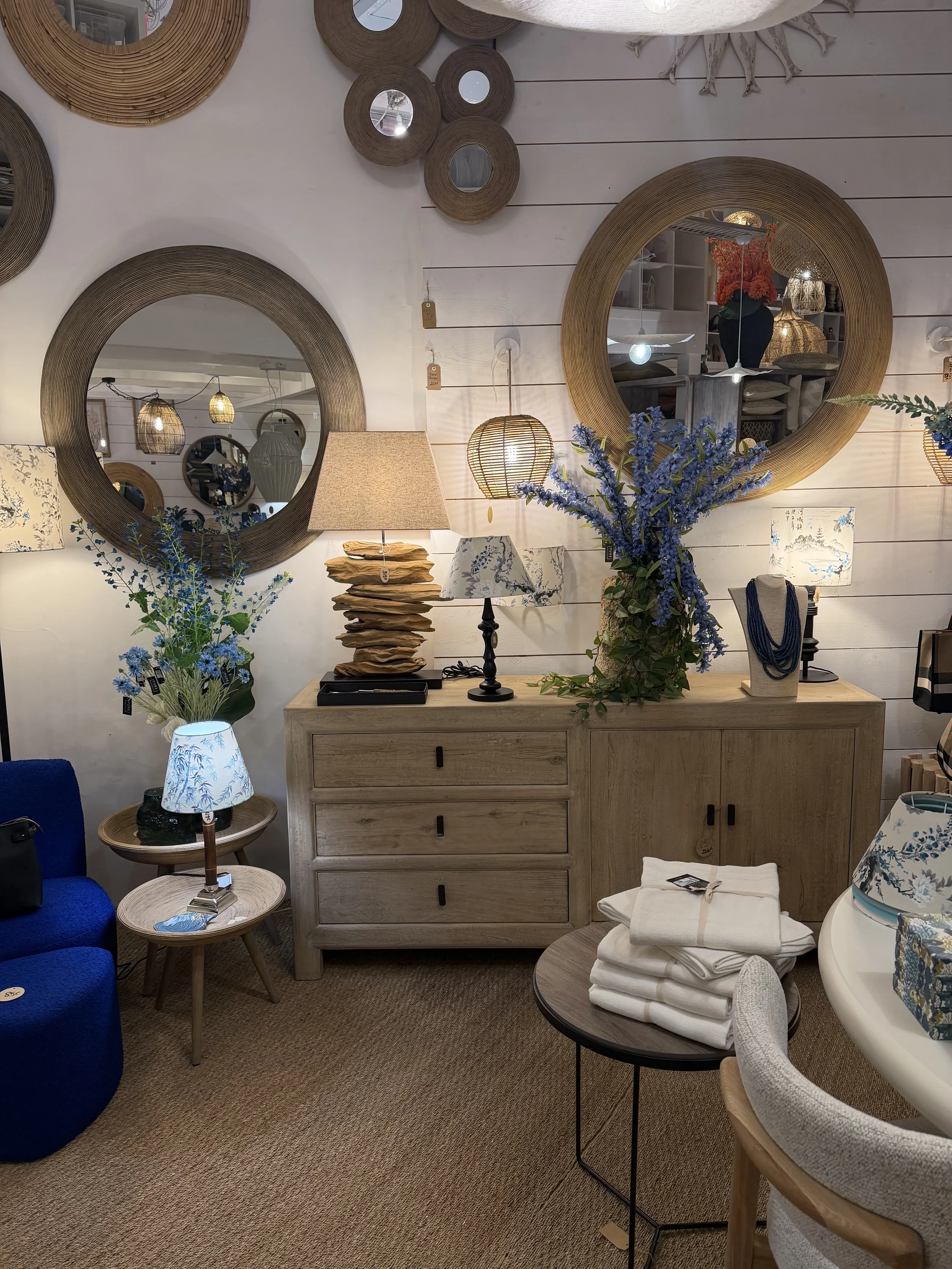 Intérieur d'une boutique de décoration avec miroirs ronds, lampes, vases de fleurs, petit mobilier en bois, et textiles blancs.