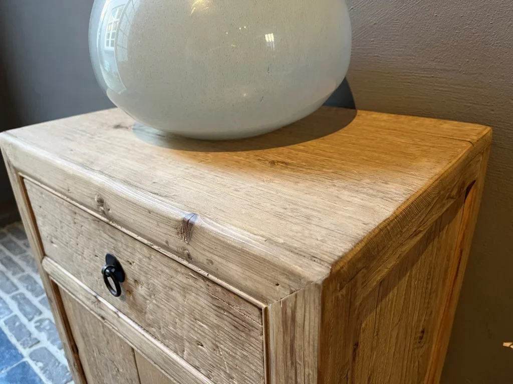 Un meuble en bois brut avec un tiroir, surmonté d'une grande vase en céramique grise, placé contre un mur gris.